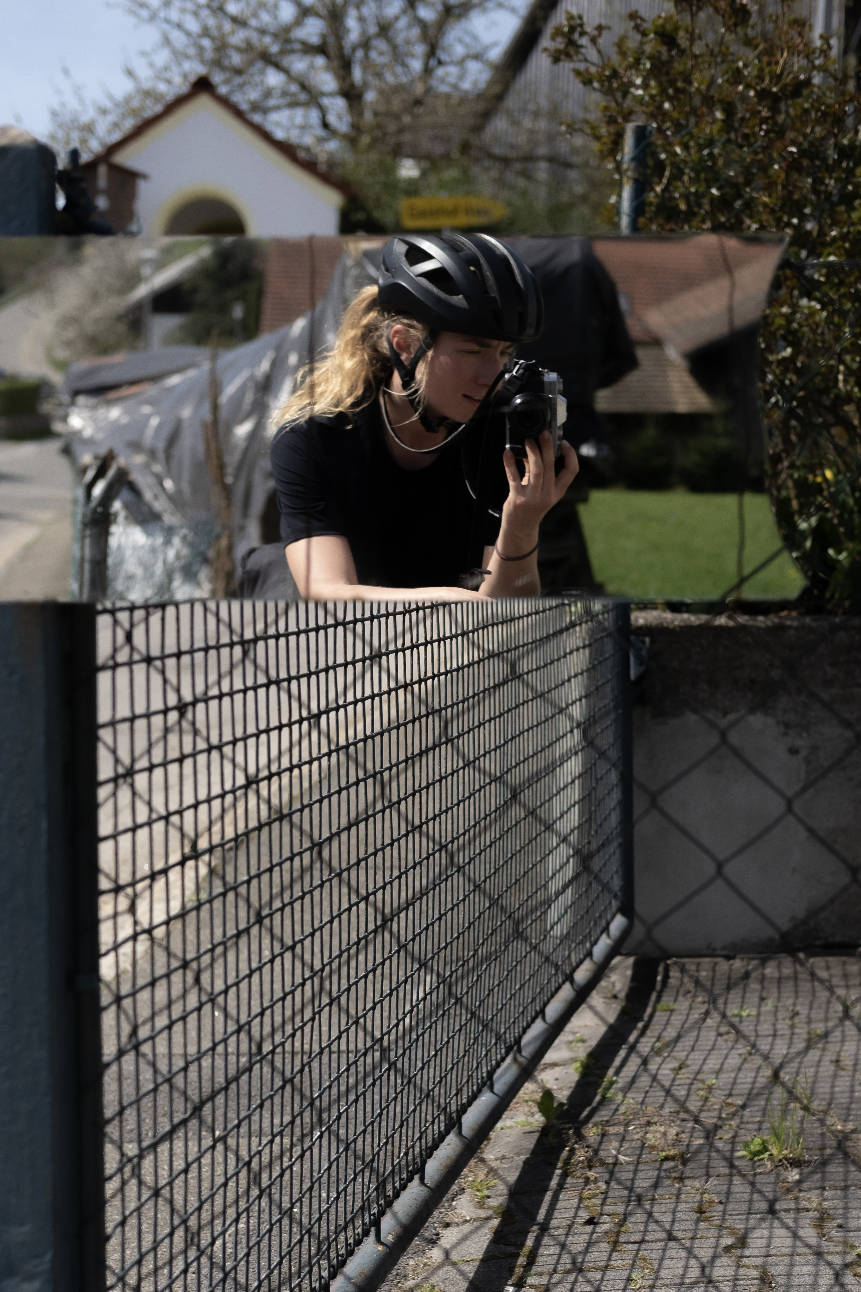 Rider in a helmet leaning on a fence, lifting a film camera to take a photo in a village