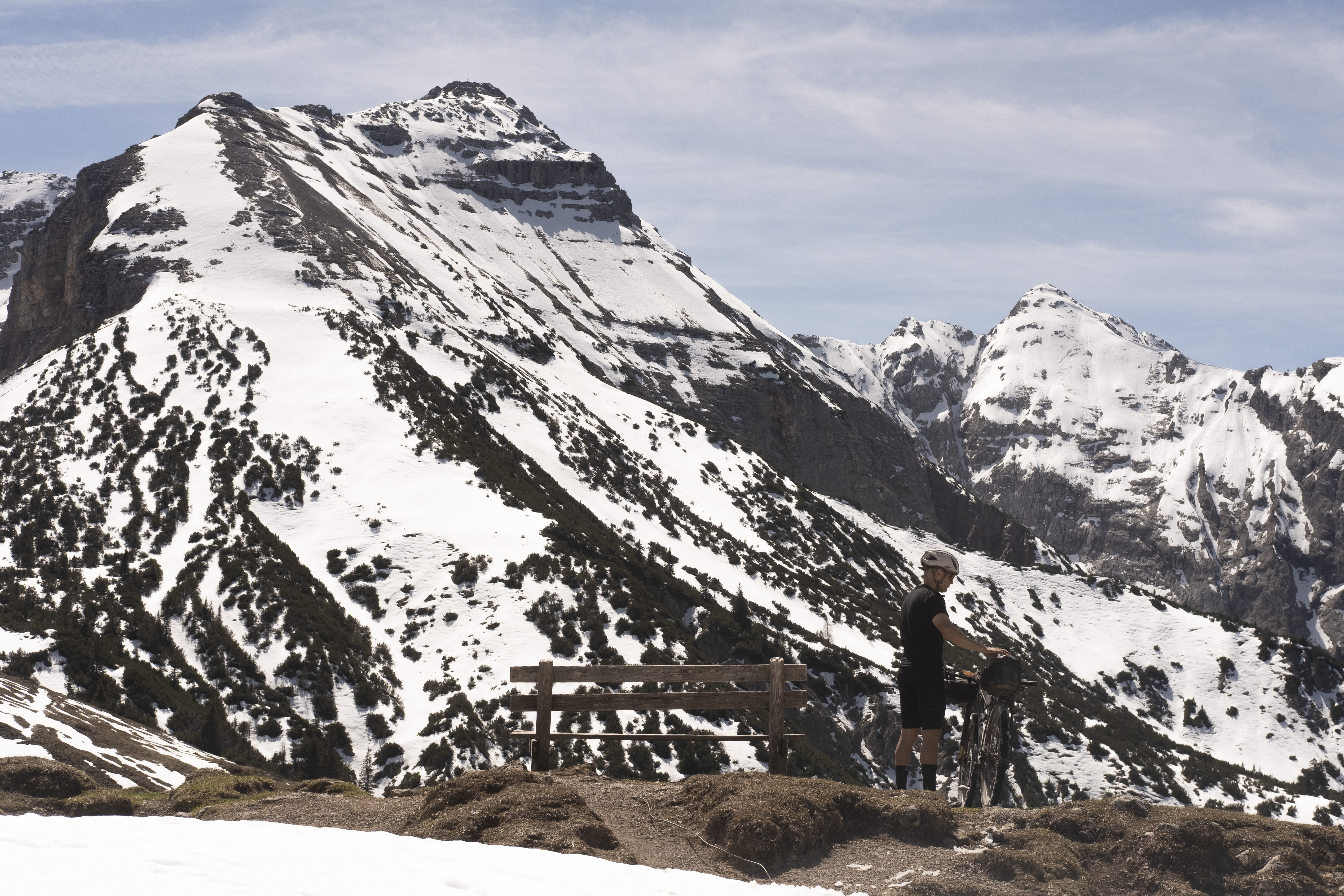 Rider standing with a bike at a wooden railing below a snow-covered limestone peak