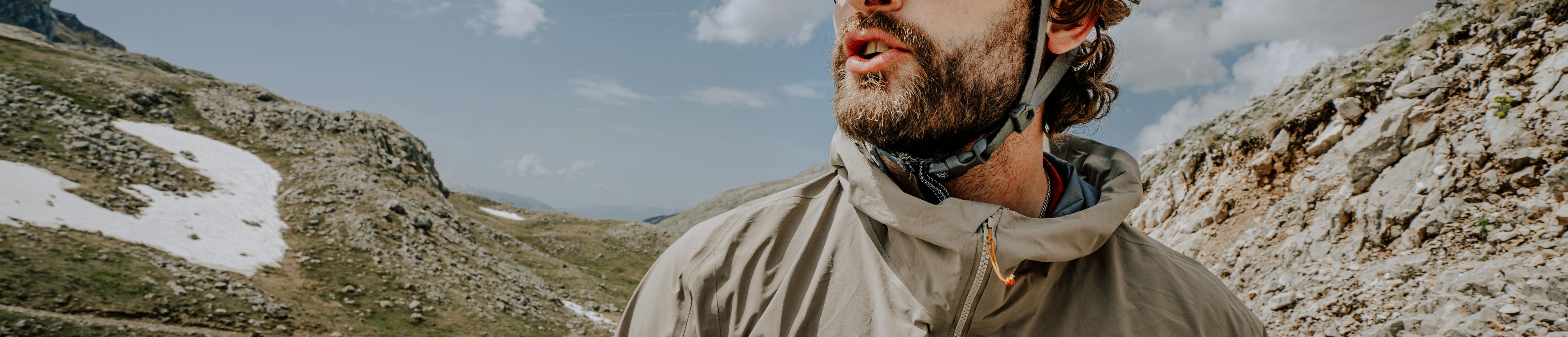 Cyclist wearing a red helmet, sunglasses, and a light 7mesh jacket stands on a mountain road, looking into the distance under a partly cloudy sky