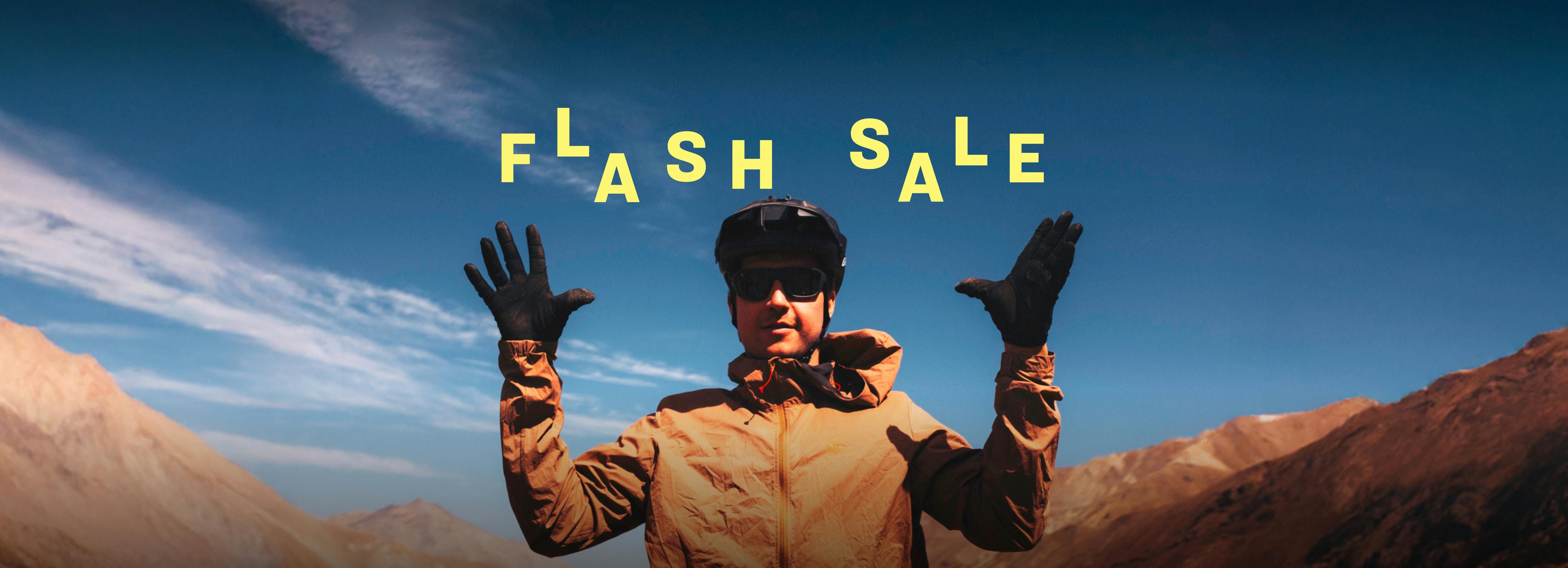 Flash Sale banner featuring a cyclist in a brown jacket and helmet raising both hands against a mountain backdrop with clear skies. Bold yellow text reads ‘FLASH SALE’ with the 7mesh logo above