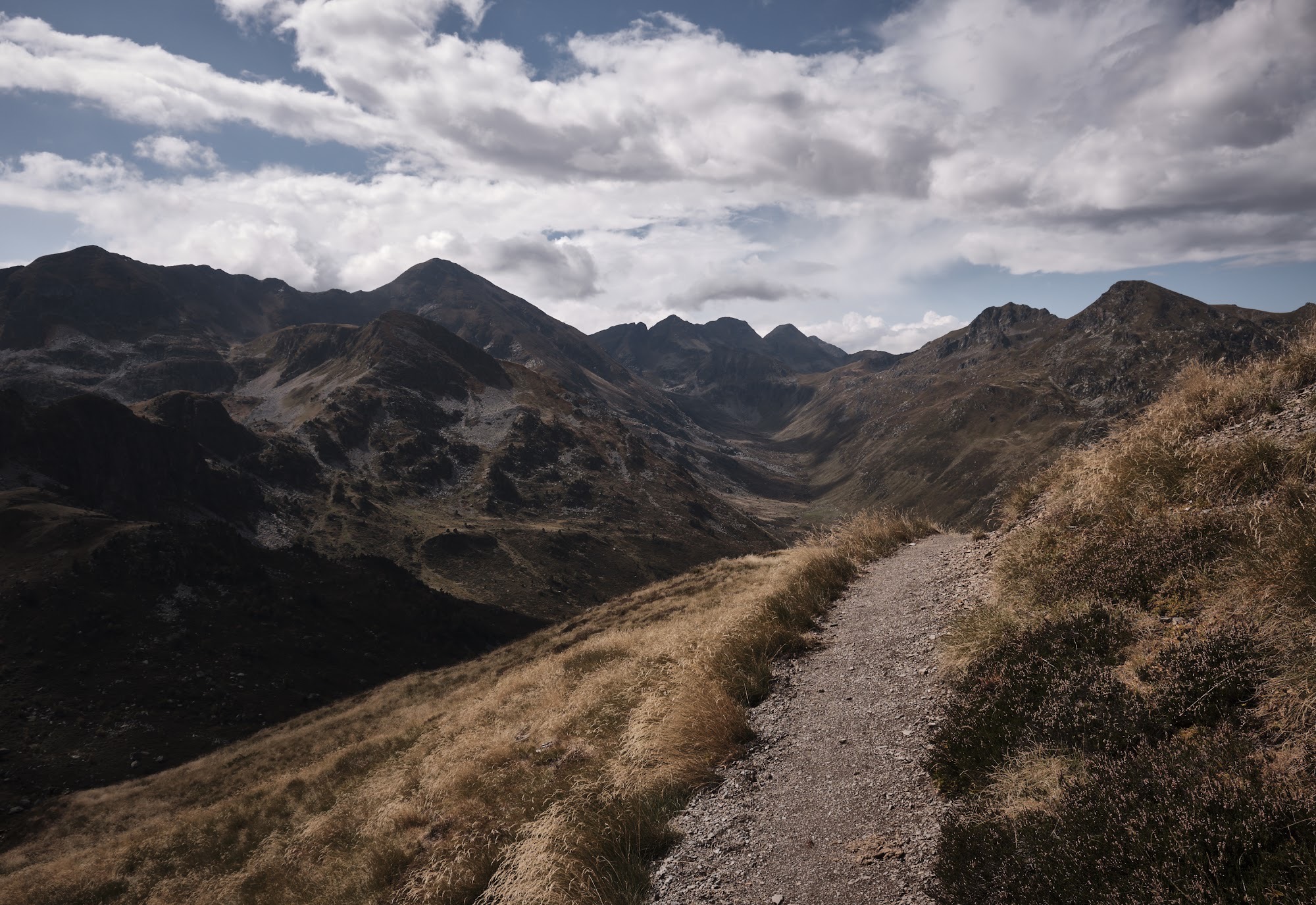Stony singletrack running along a ridge toward jagged grey peaks under broken cloud