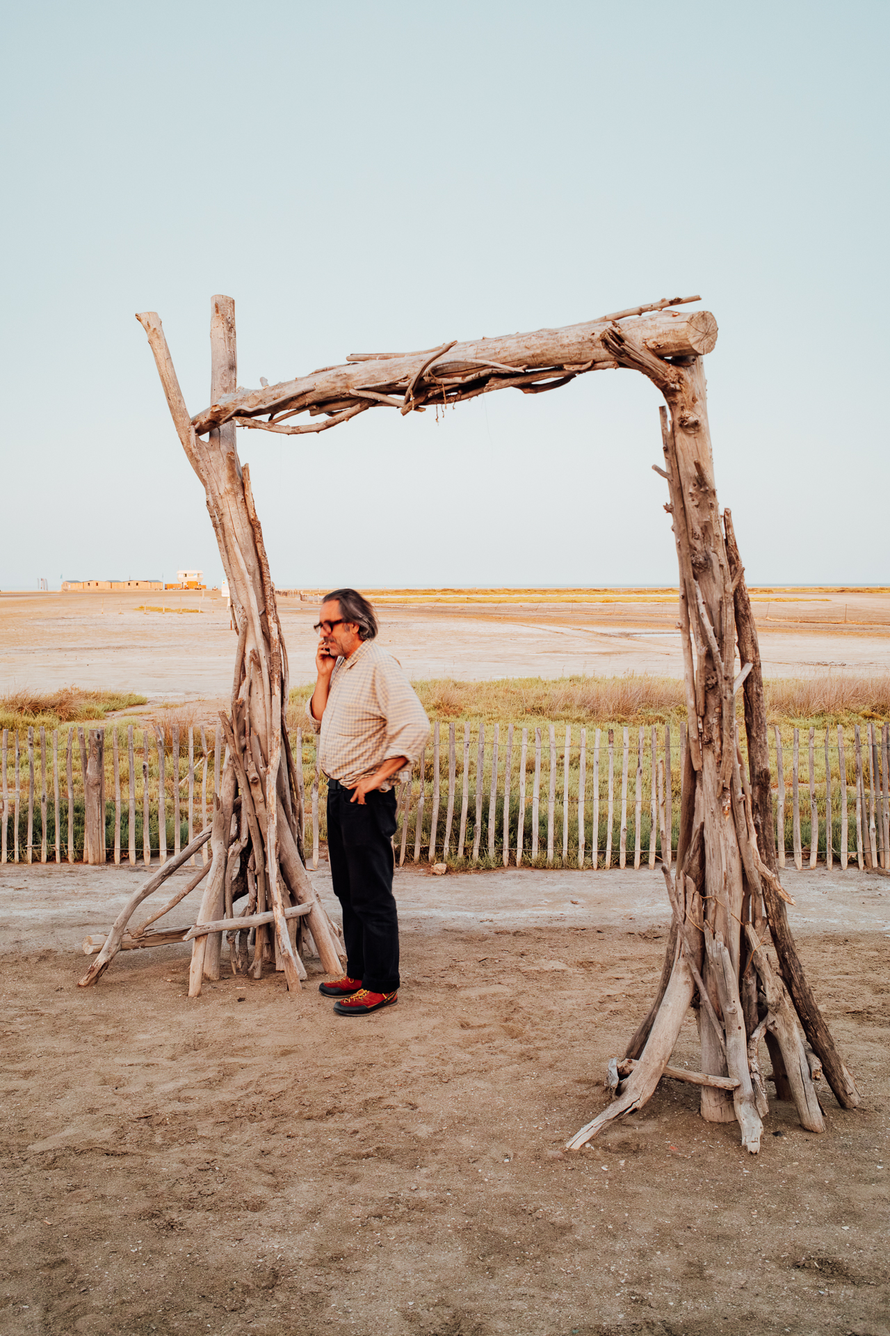 Man with long grey hair on the phone beside driftwood tripods on a flat sandy shore