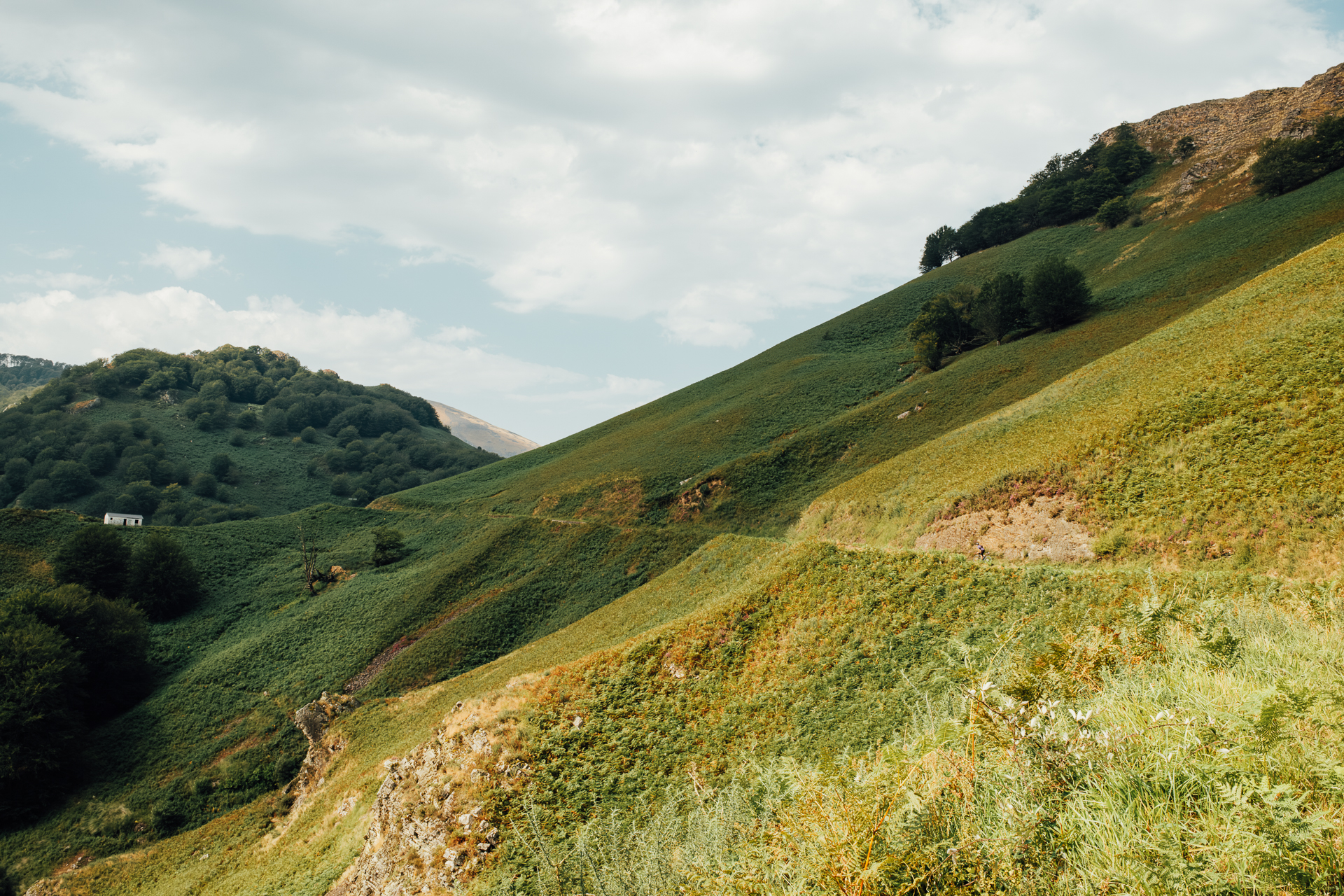 Steep green hillside with a faint traversing path and a small white building in the valley below