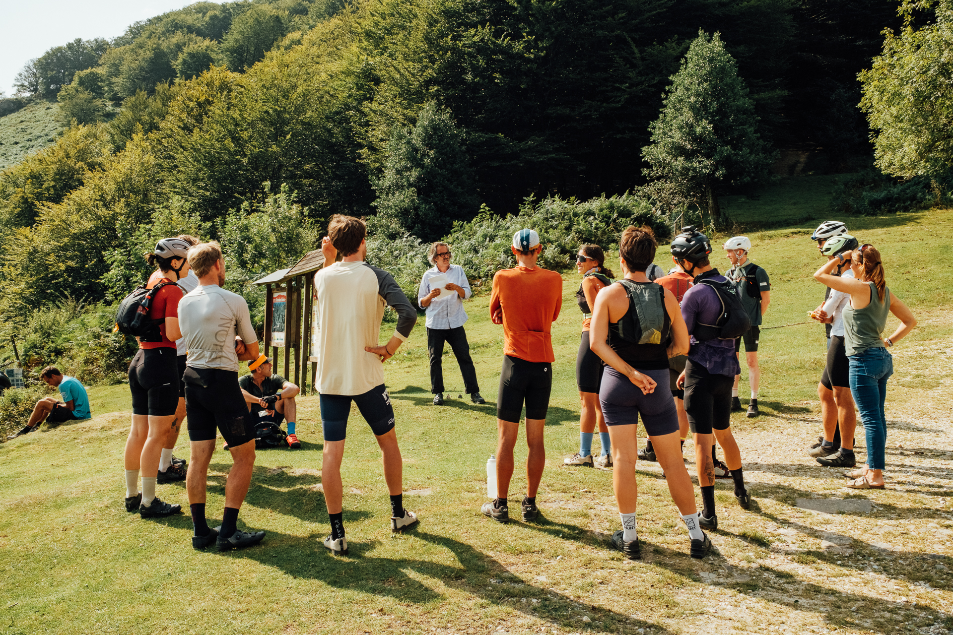 Cyclists in shorts and helmets standing in a grassy clearing, listening to a man in a white shirt