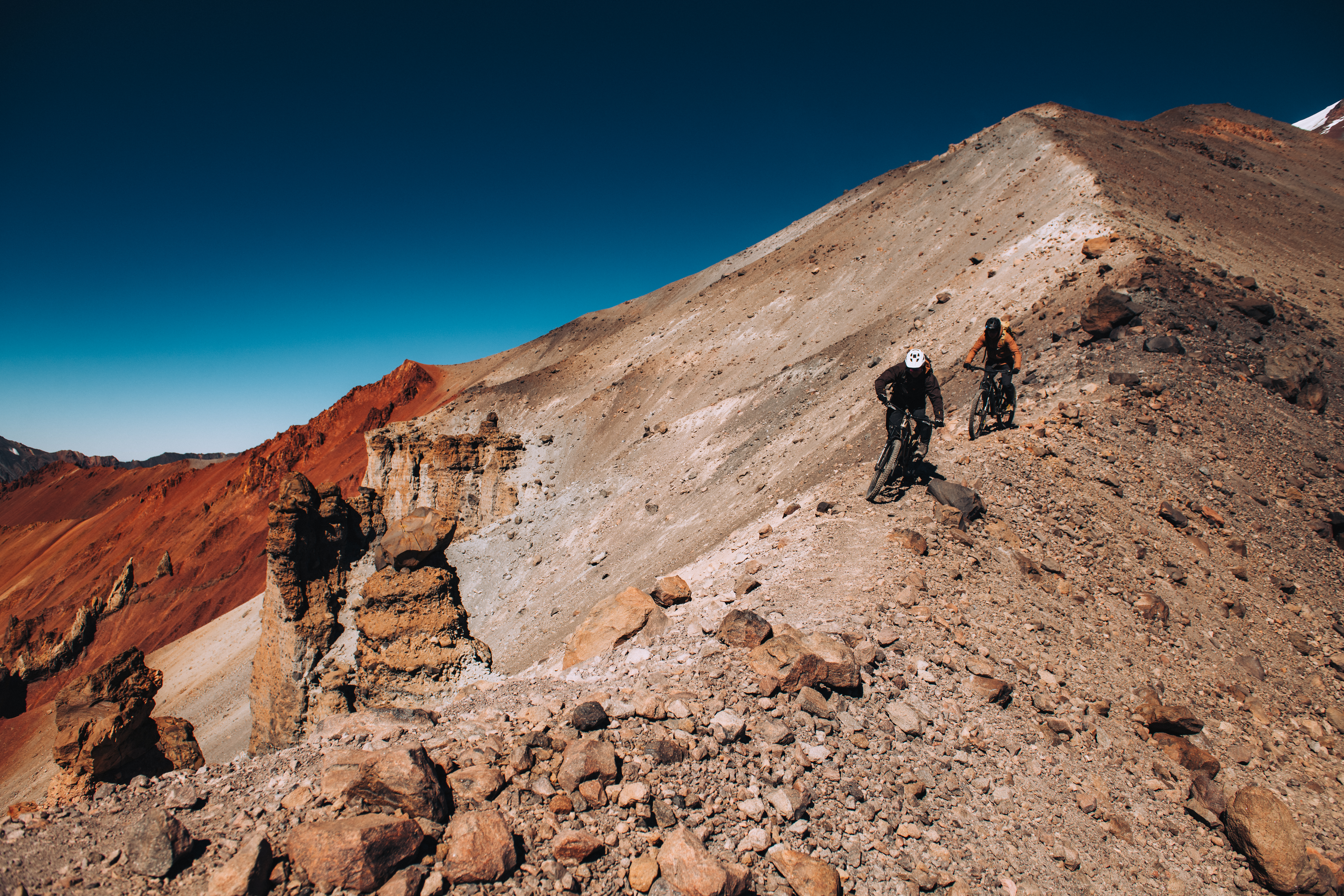 Two riders on a red and grey volcanic ridge under deep blue sky