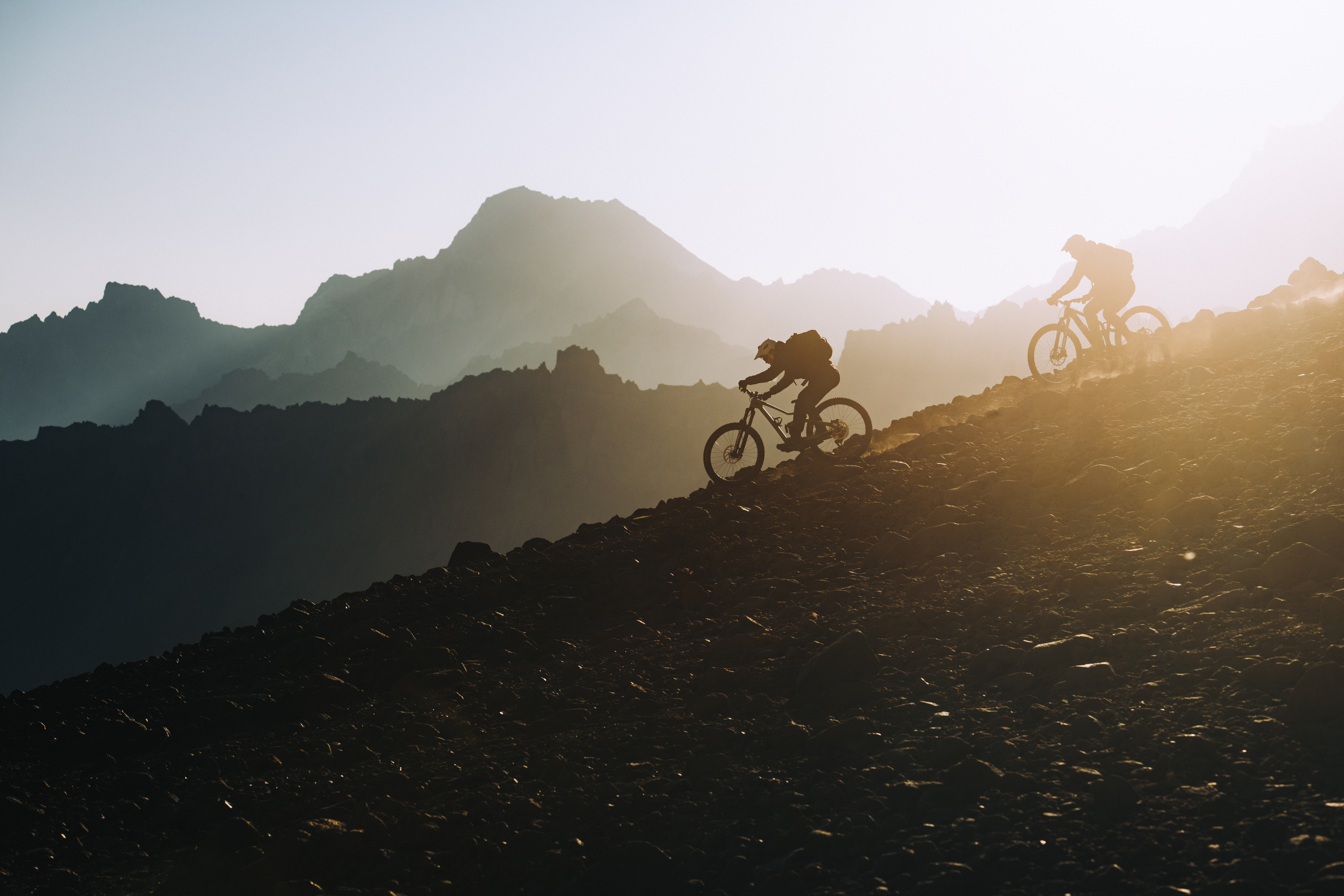 Two riders silhouetted descending a dark slope into low sun