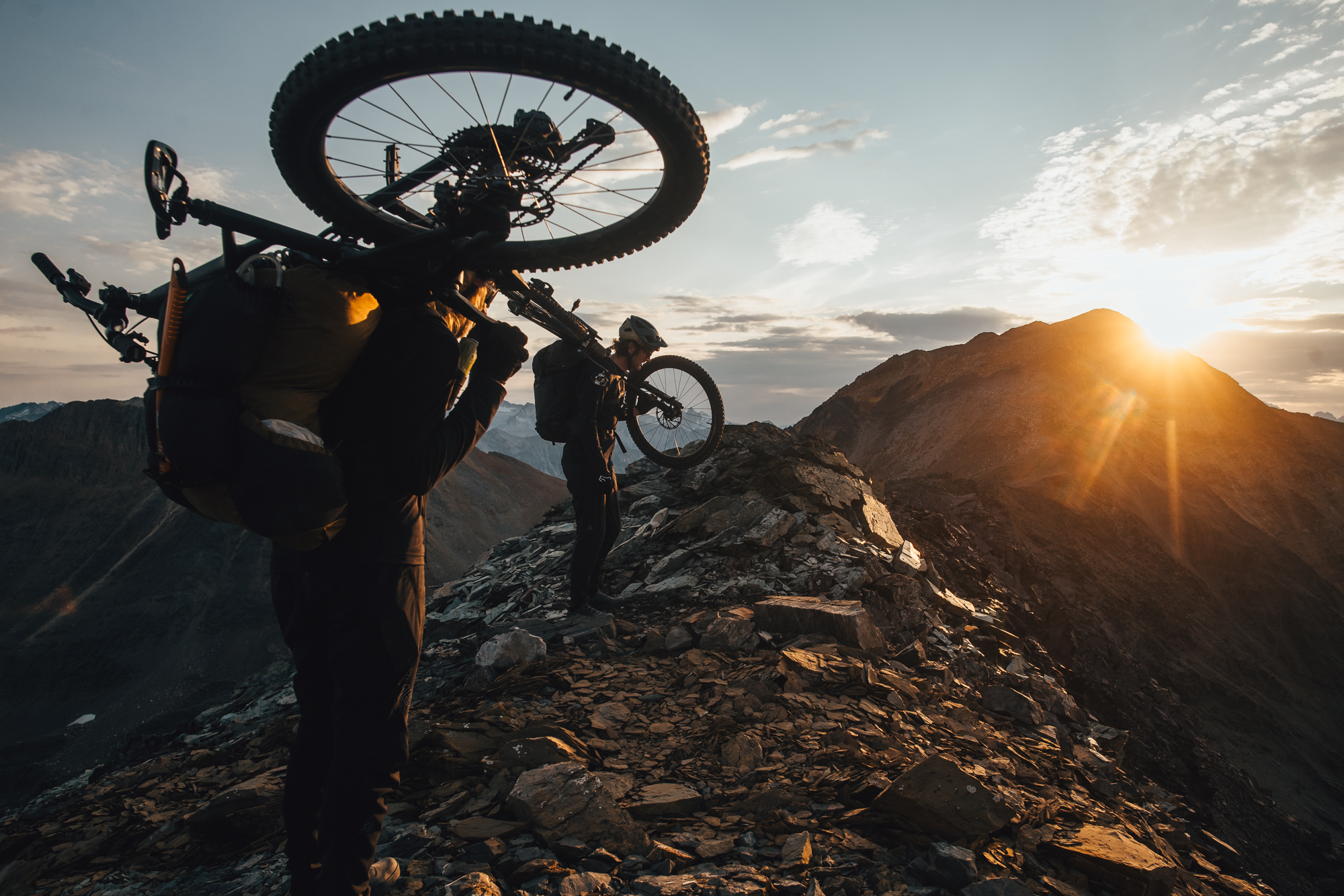 Rider carrying a loaded bike on their shoulder into low sun on a ridge