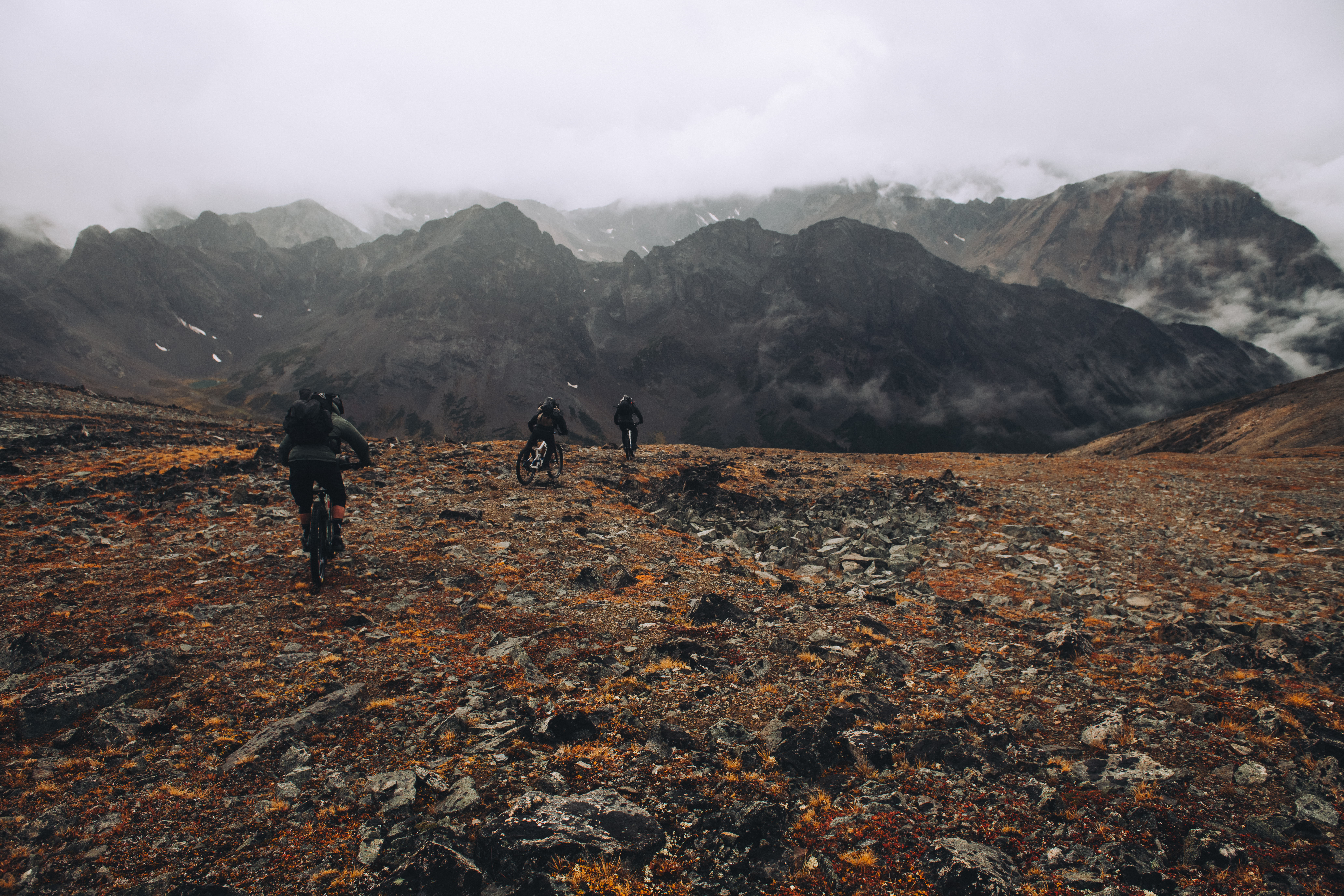 Three riders pushing bikes up an open scree slope below cloud