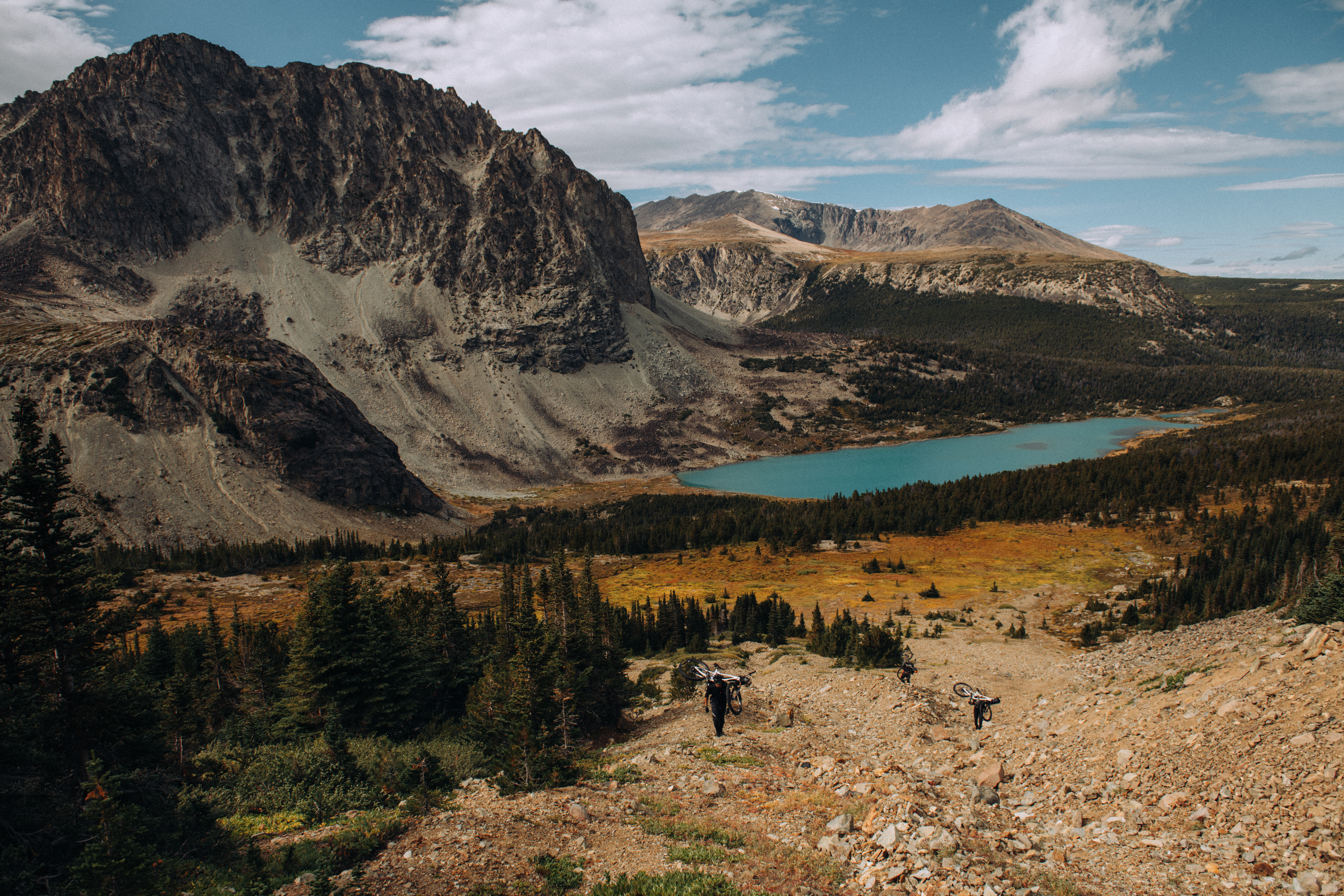 Riders on a rocky trail above a turquoise lake and larch meadow