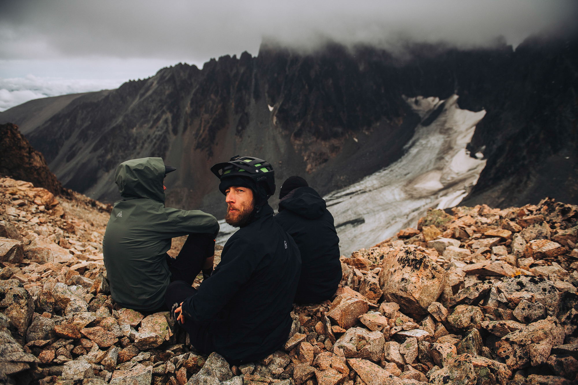 Three riders sitting on scree looking out at cloud-covered peaks