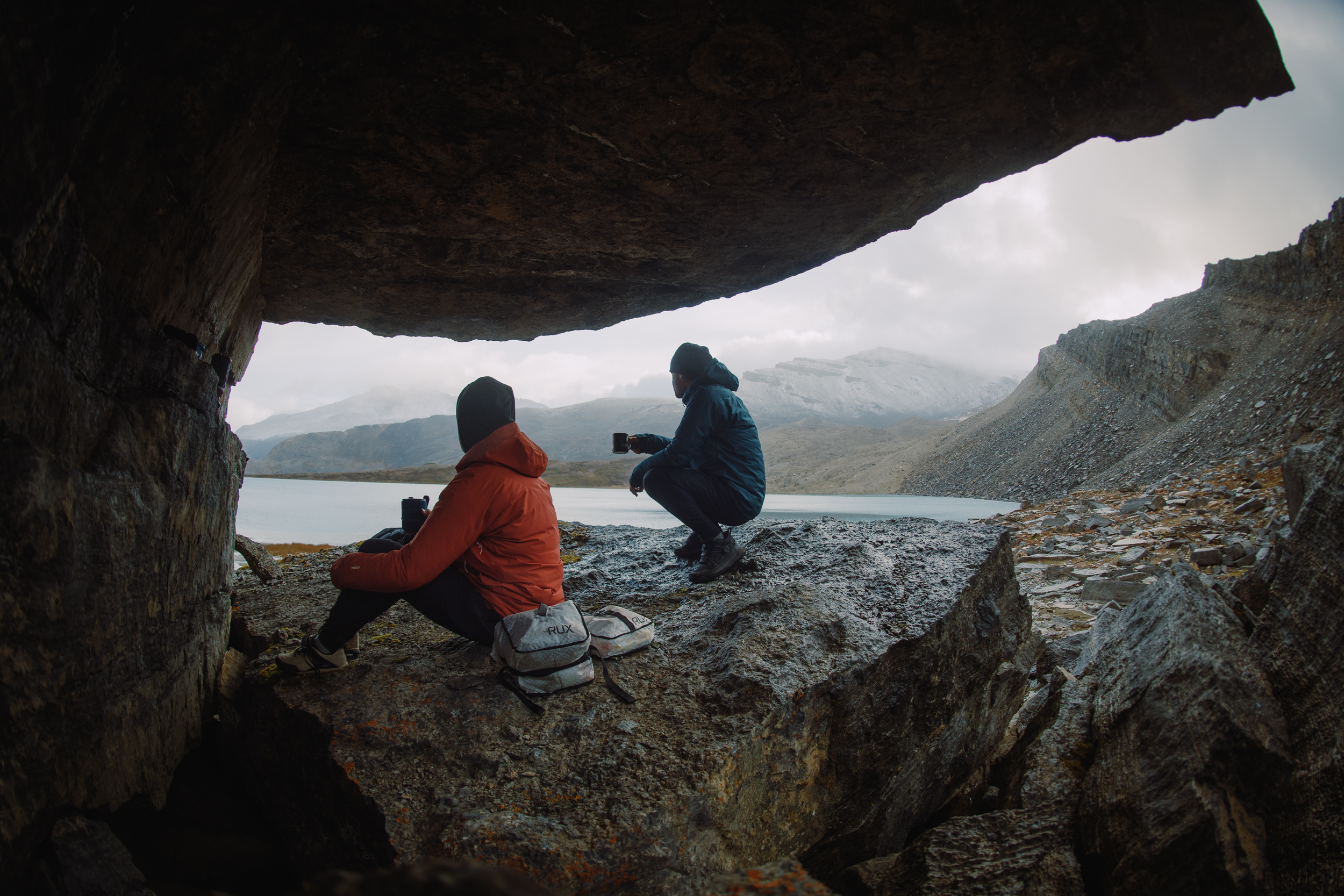 Two riders drinking from mugs under a rock overhang above a lake