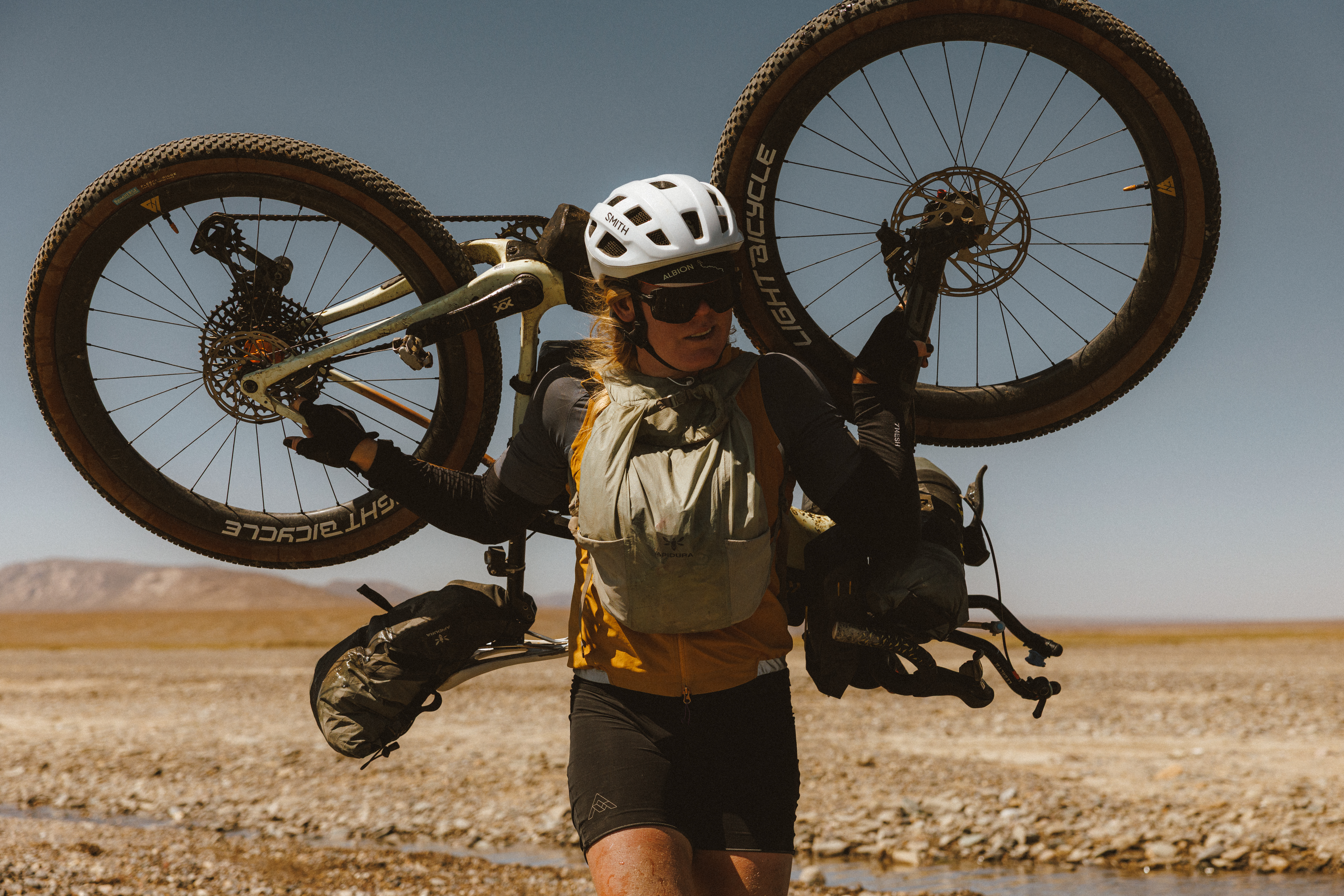 Rider carrying a loaded bike on her shoulder across an open gravel plain