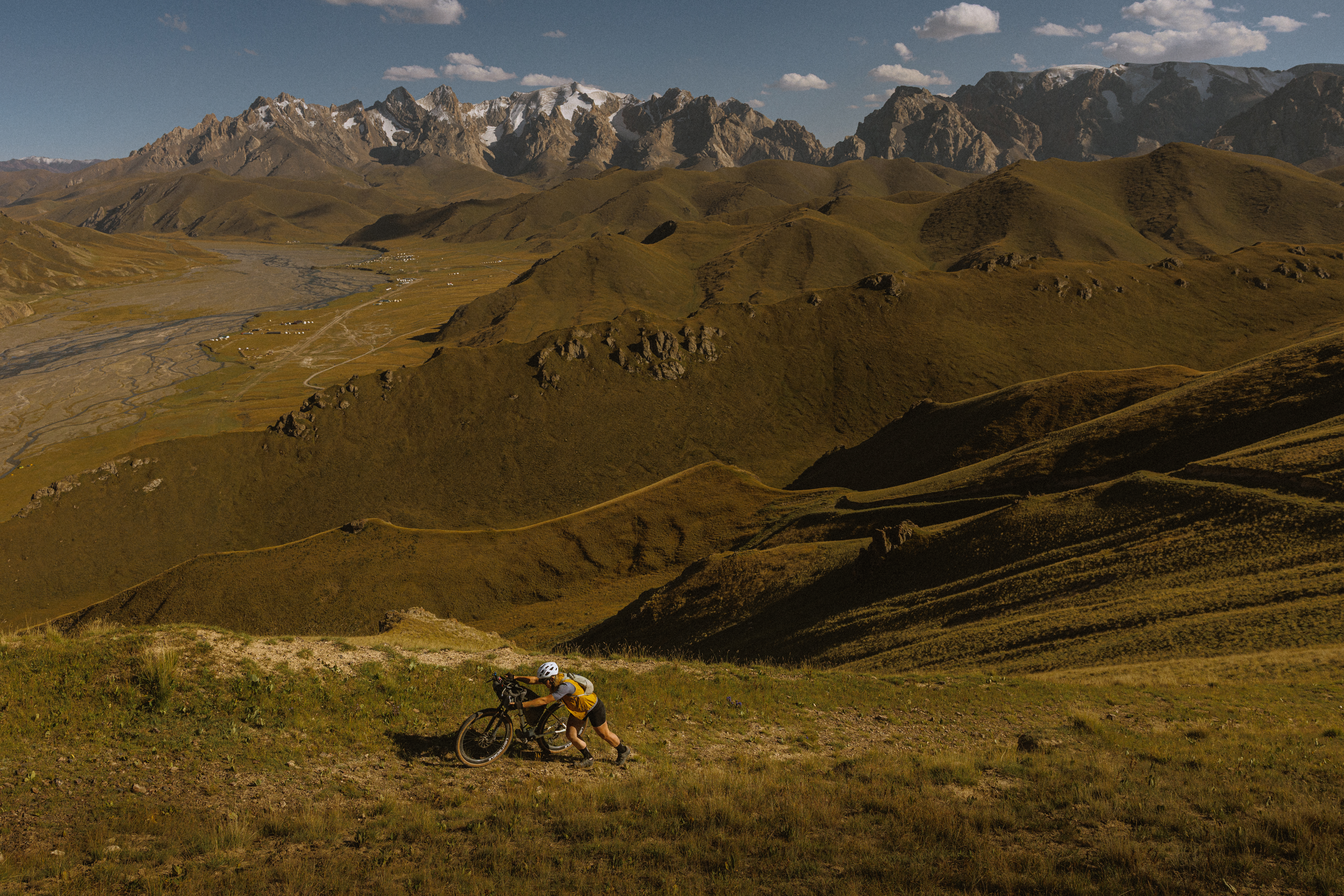 Tiny rider on a track below huge folded hills and a jagged mountain range