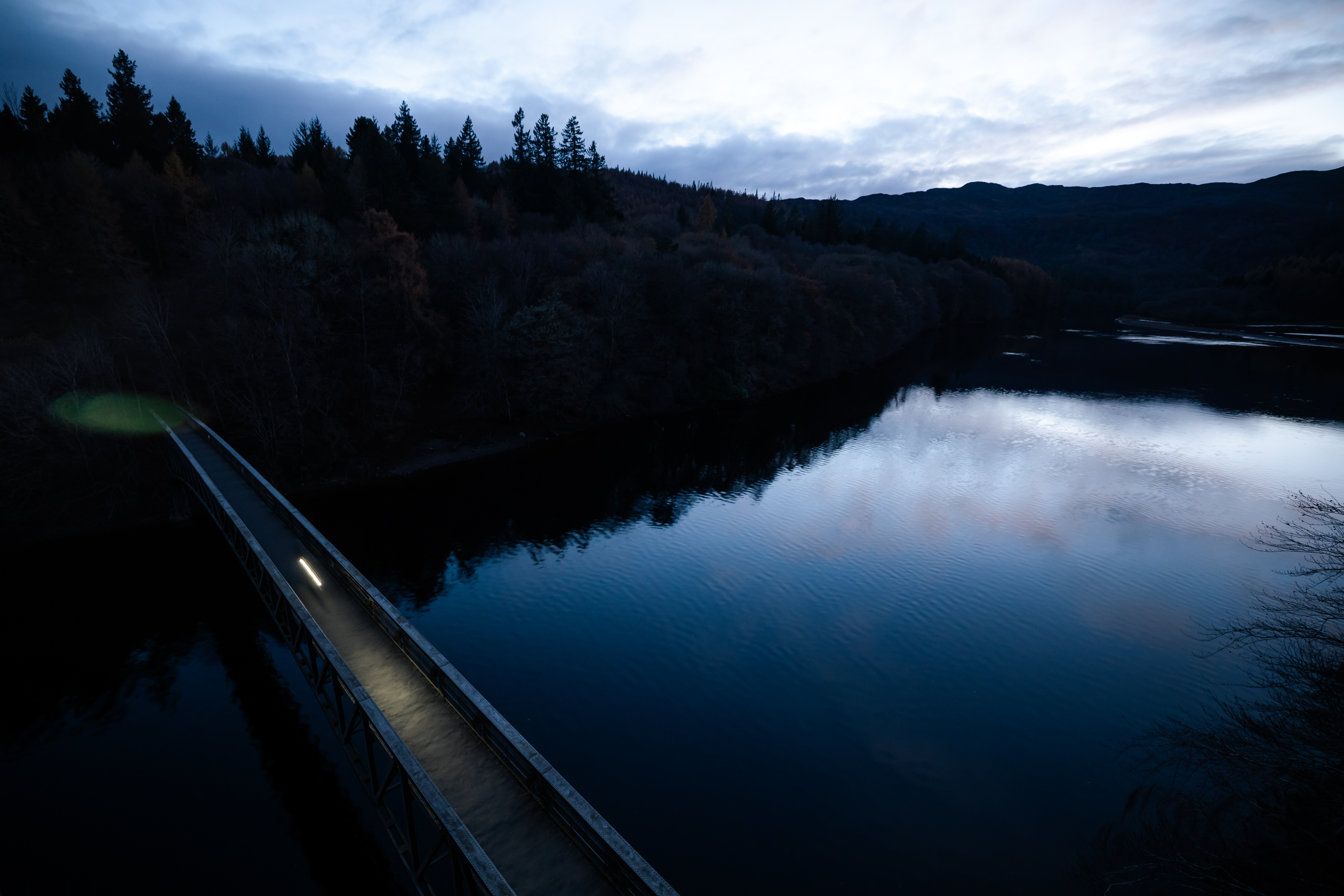 Lit bridge railing over still water at dusk below dark forested hills