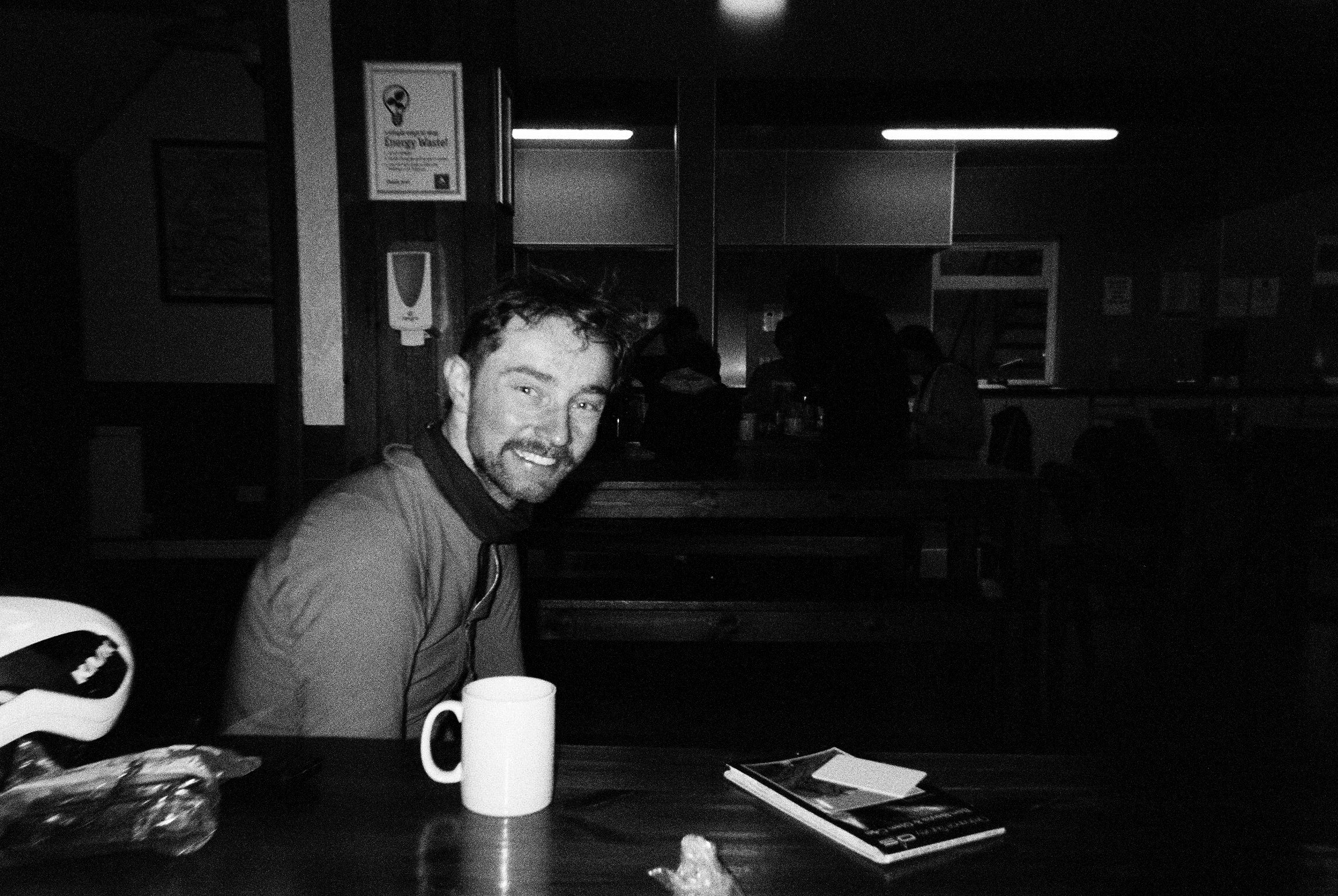 Rider smiling over a mug at a table in a lit hall, black and white