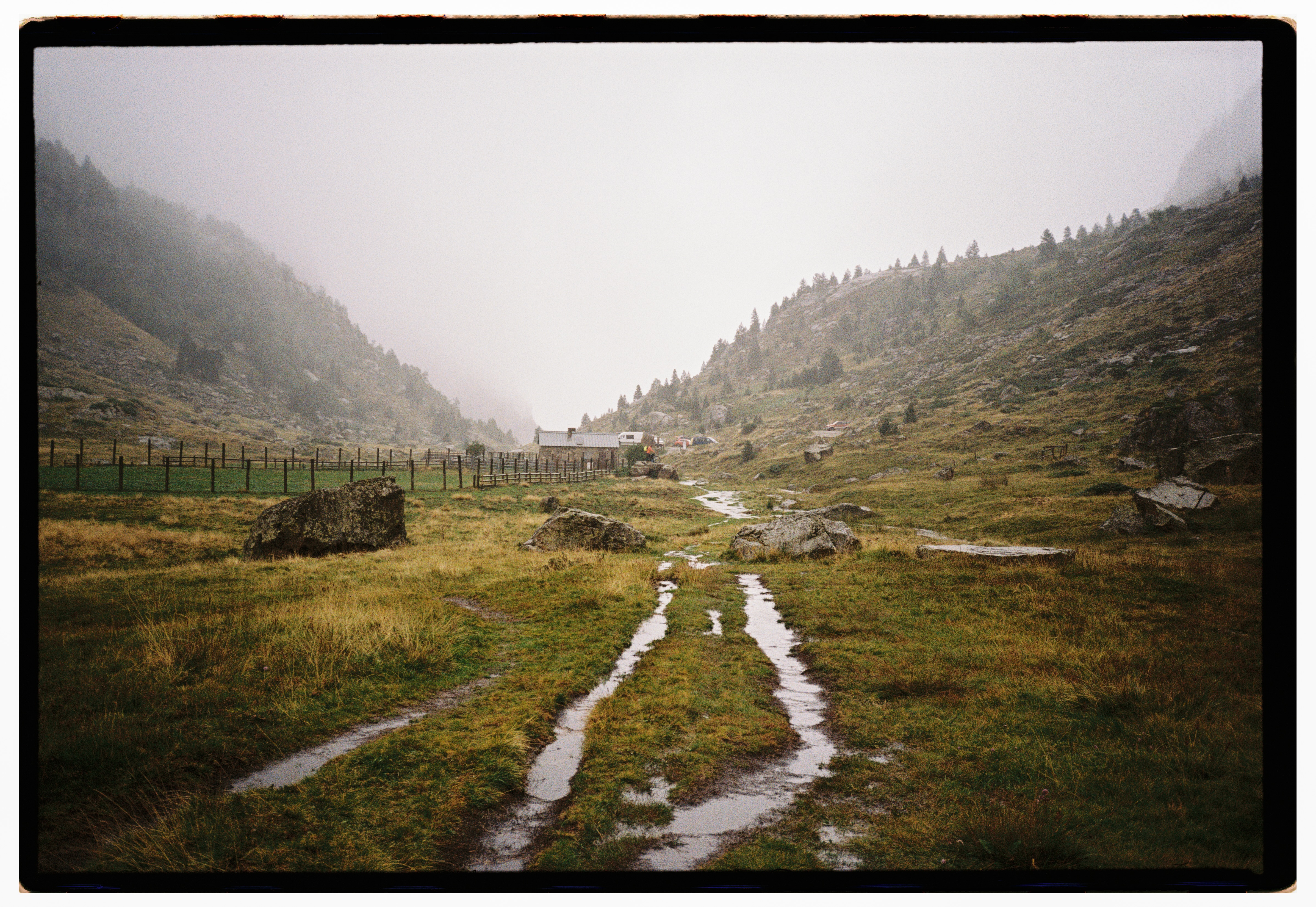 Wet double-track running down a misty green valley toward farm buildings