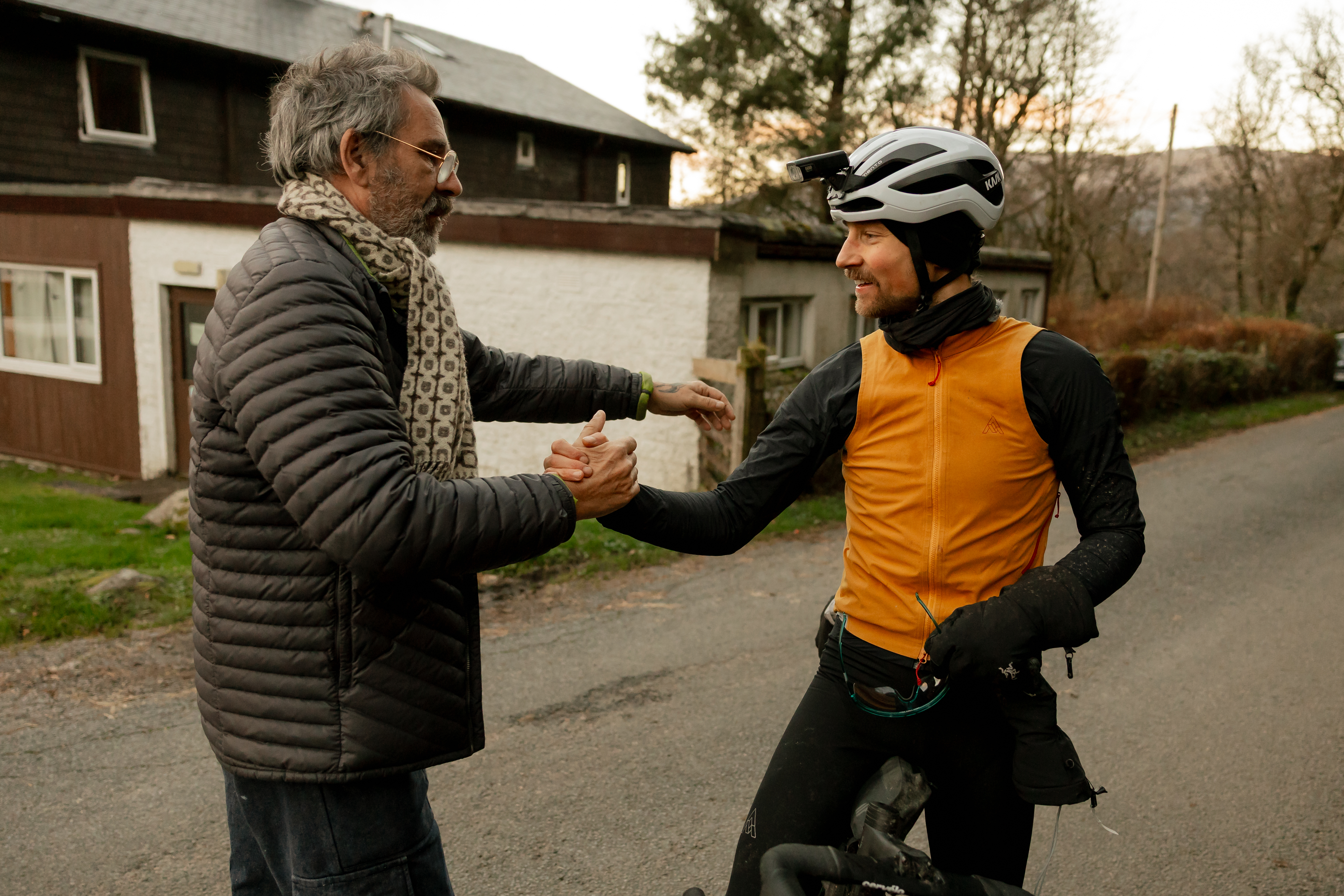 Rider shaking hands with an older man on a road outside a stone building