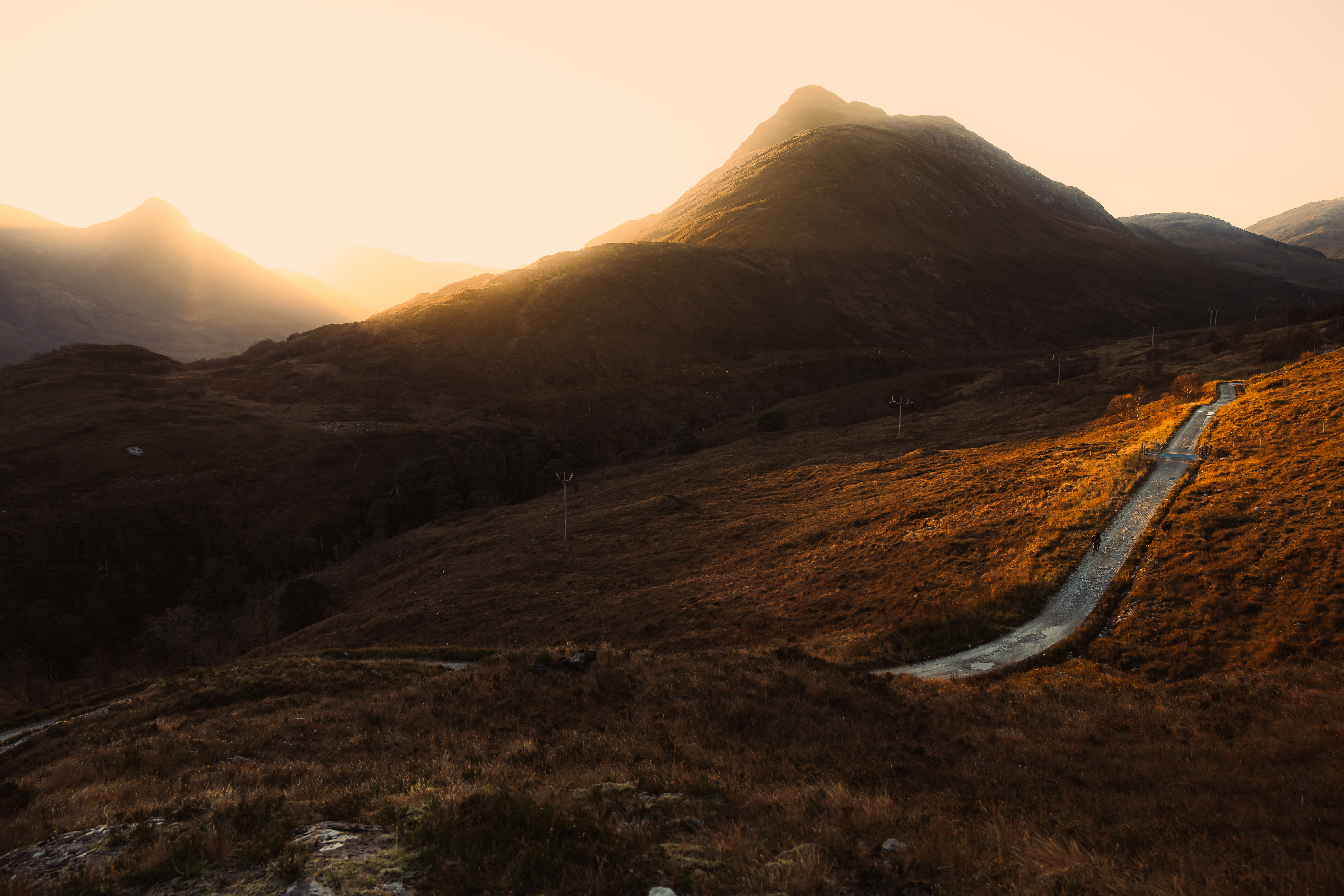 Single-track road winding up a sunlit hillside toward a dark peak at dawn
