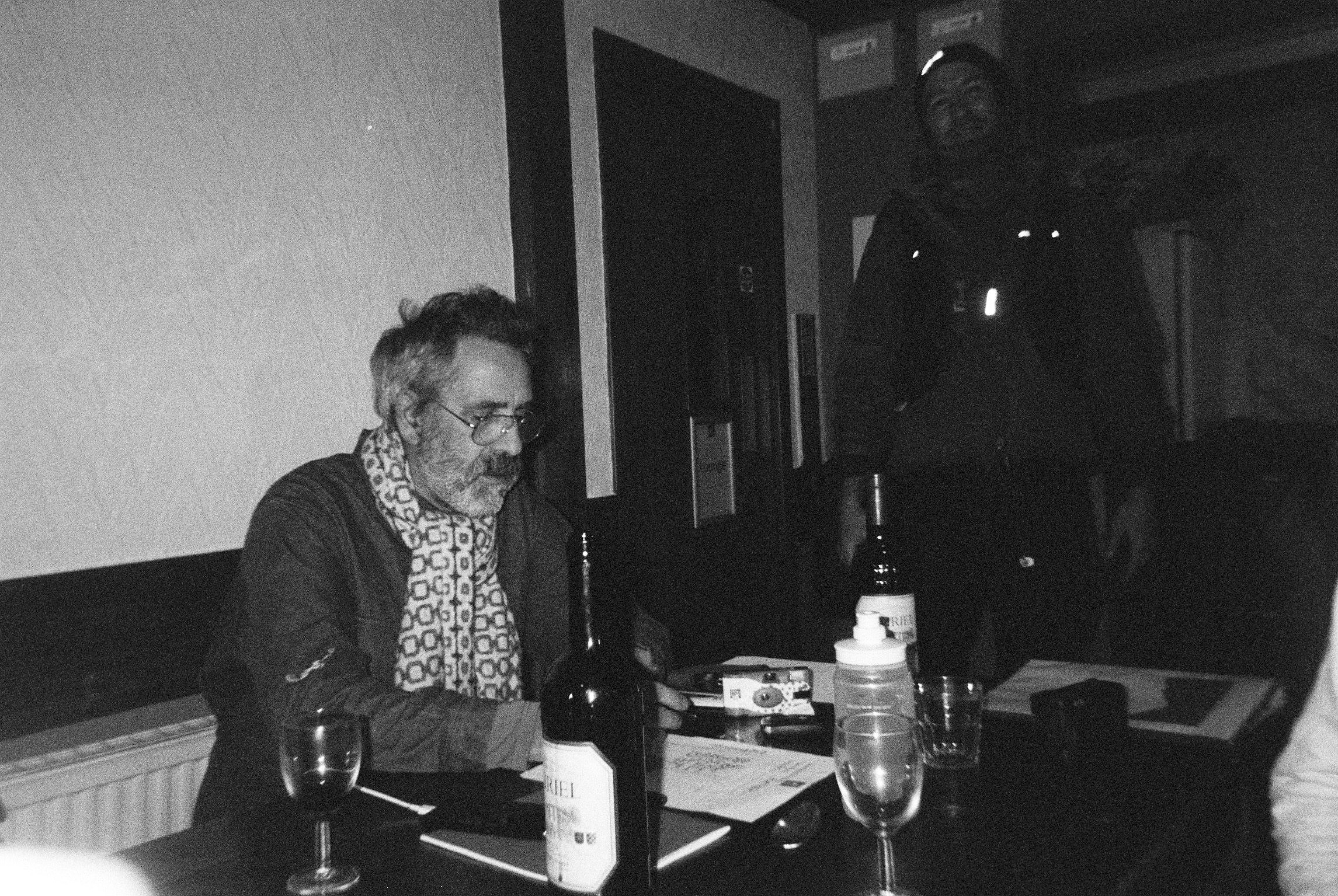 Man in a scarf at a table of wine bottles with a rider standing behind