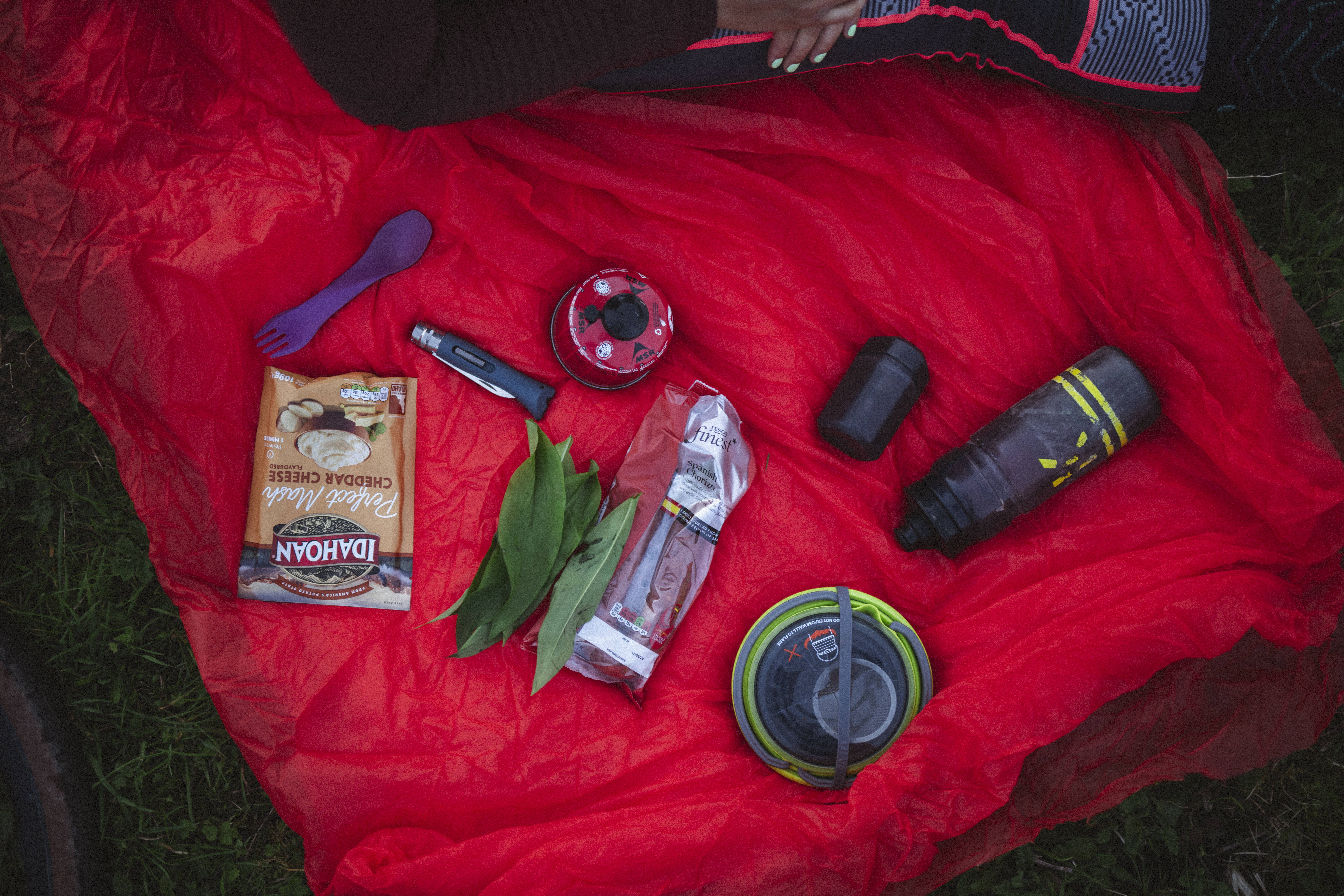 Camp kit laid out on a red bivvy: instant mash, stove, spork and flask
