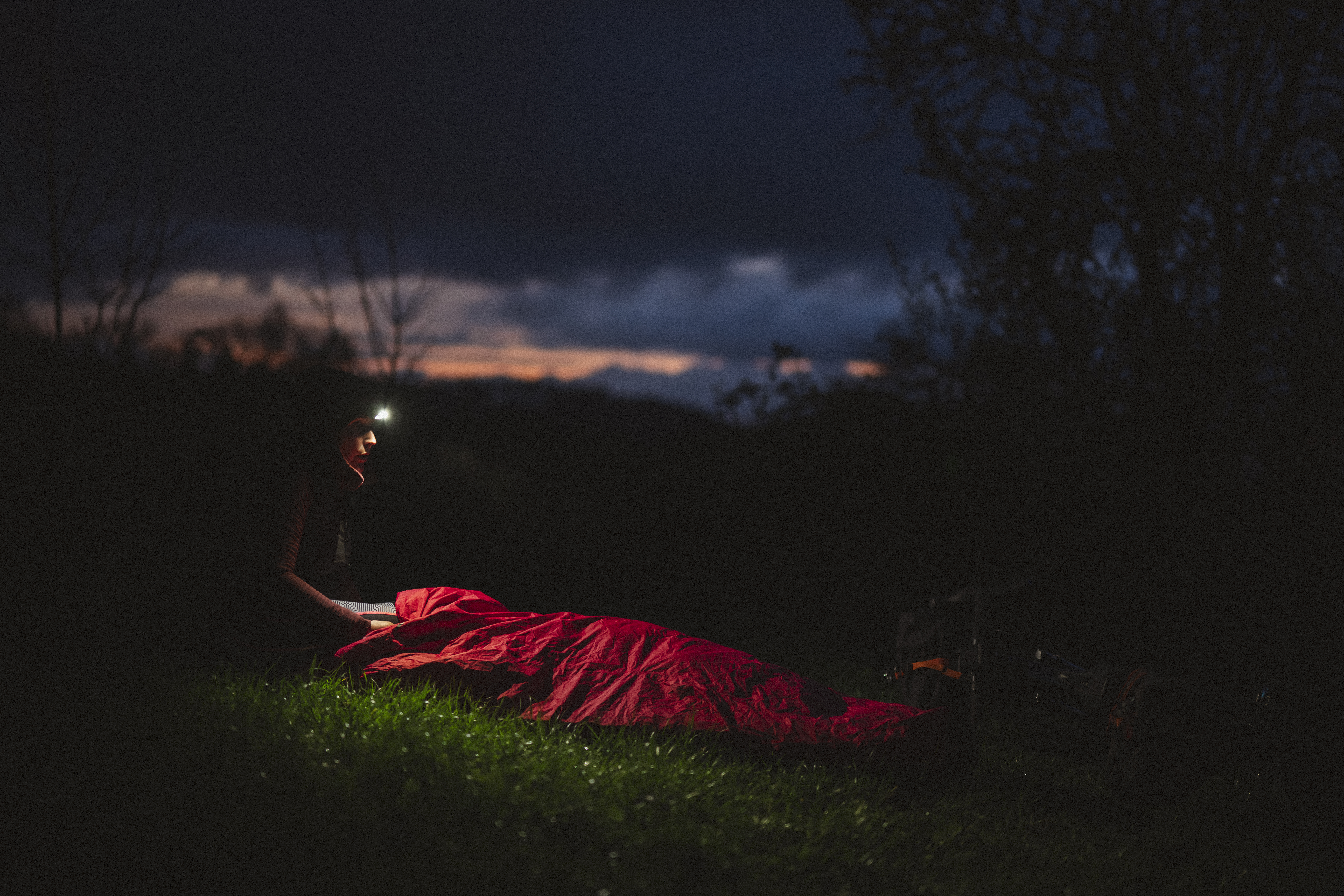 Rider in a head torch beside a red bivvy on grass at dusk