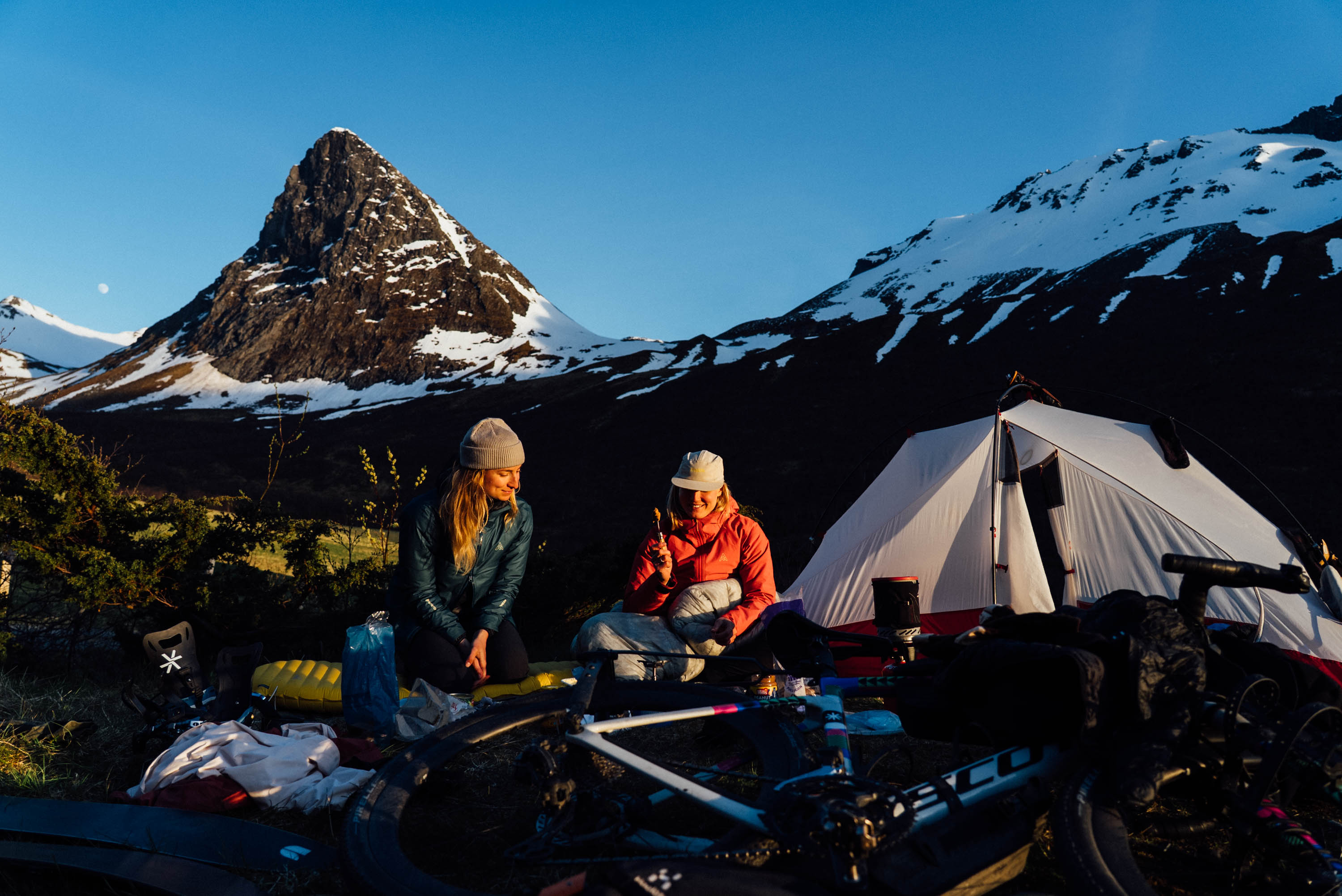 Two riders at a tent camp below a snow-streaked peak at dusk