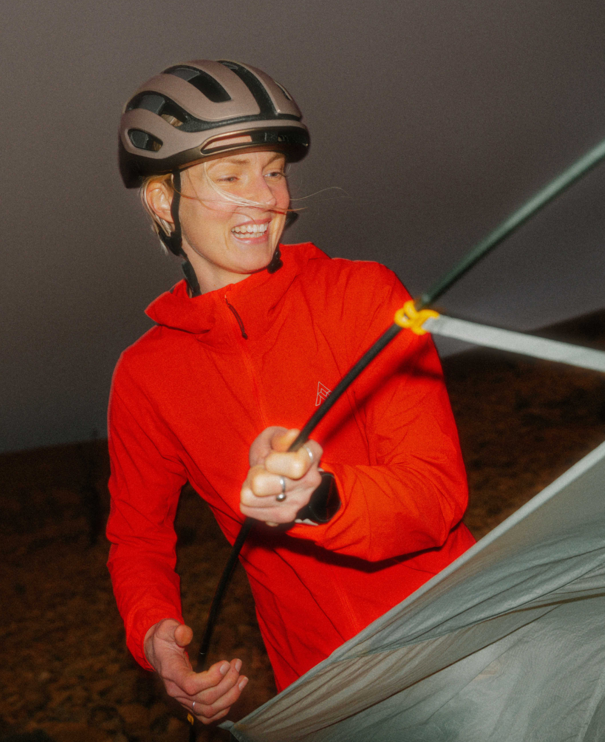 A cyclist wearing a matte brown helmet and the vibrant red 7mesh Northwoods Jacket smiles while setting up a tent at dusk. The jacket’s lightweight, technical fabric is highlighted as she holds a tent pole, emphasizing its utility for bikepacking and transitional light.