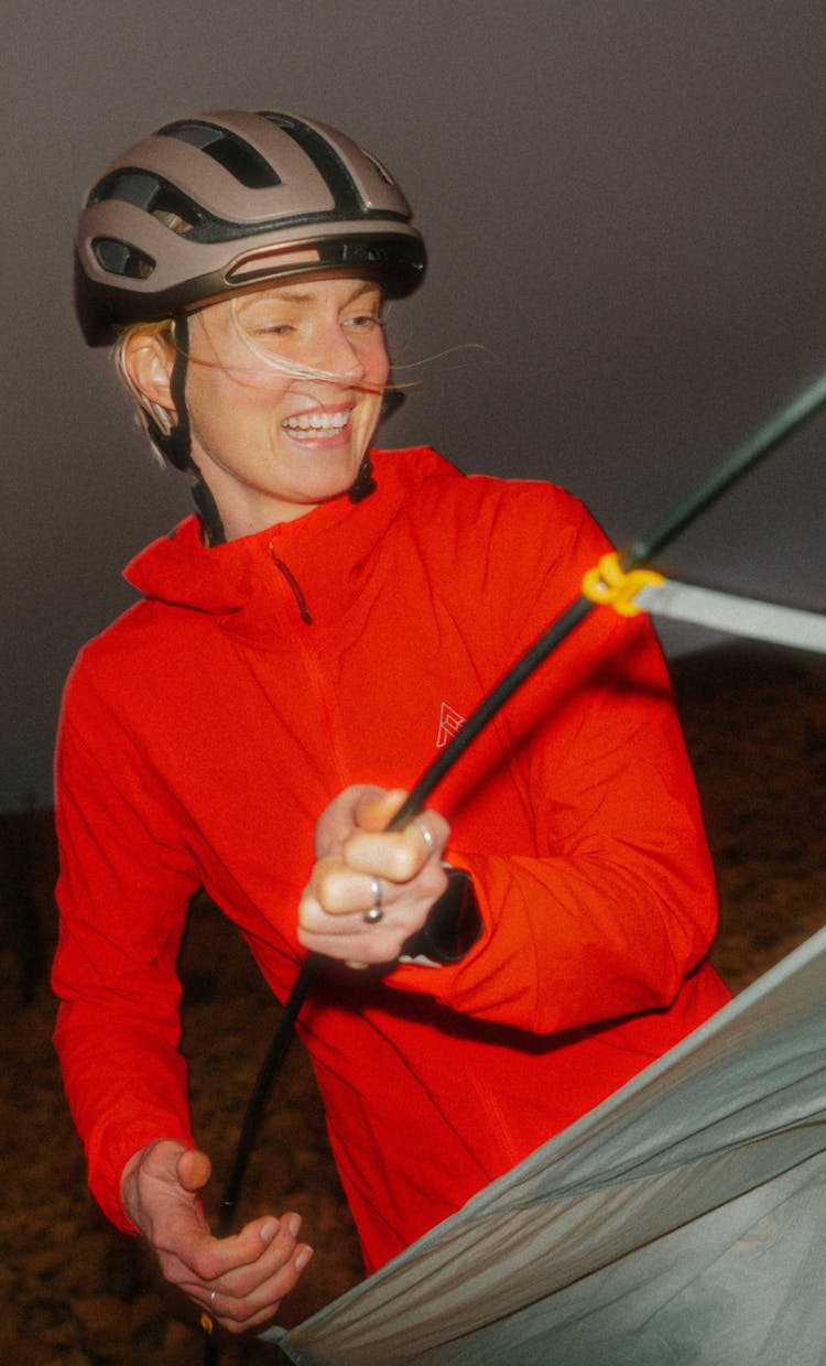 A cyclist wearing a matte brown helmet and the vibrant red 7mesh Northwoods Jacket smiles while setting up a tent at dusk. The jacket’s lightweight, technical fabric is highlighted as she holds a tent pole, emphasizing its utility for bikepacking and transitional light.