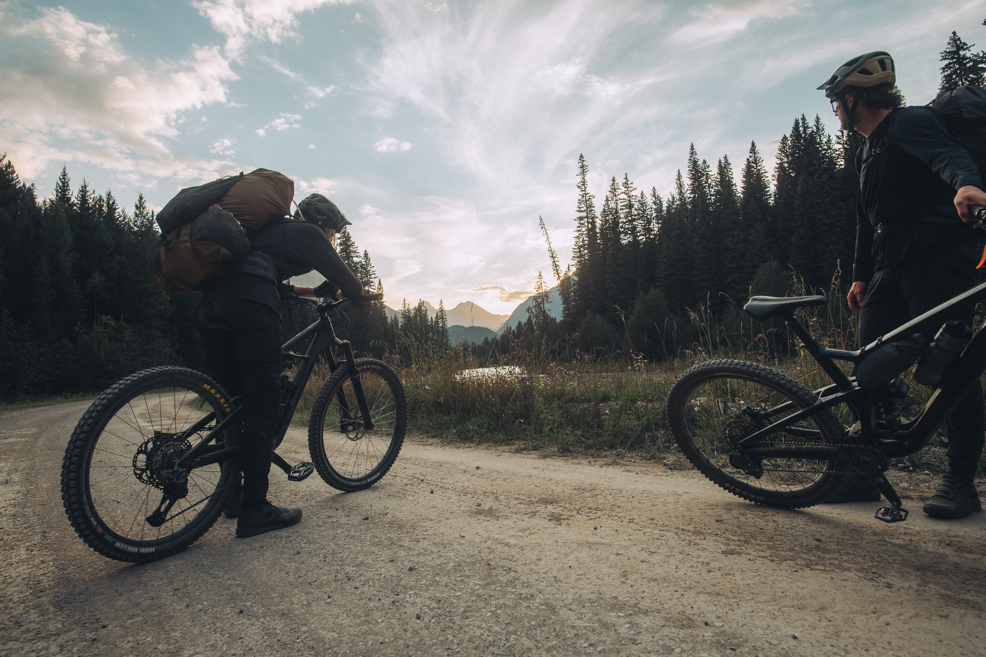 Two riders pushing bikes along a gravel road at dusk