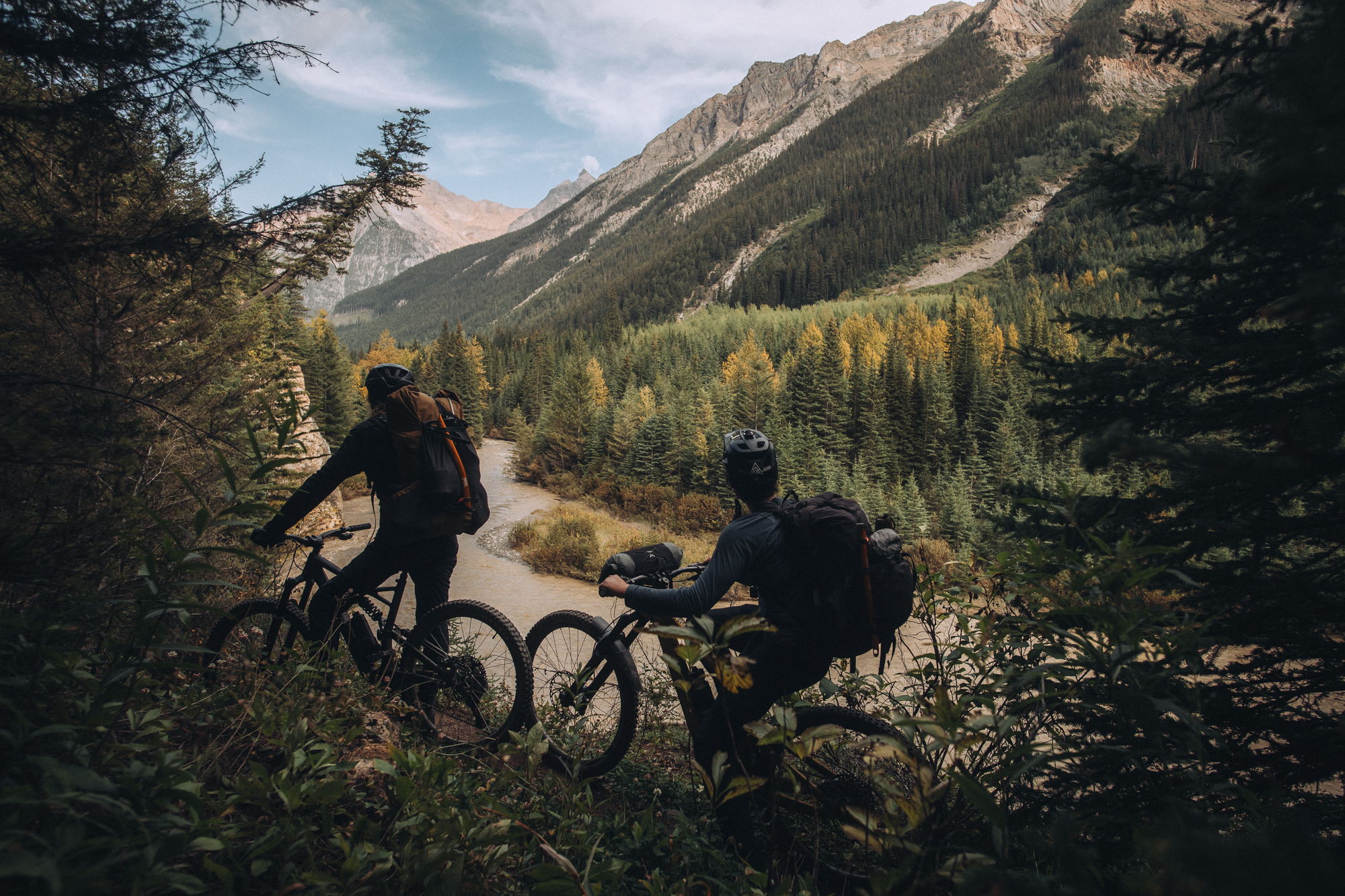 Two riders on a narrow trail above a forested valley and river