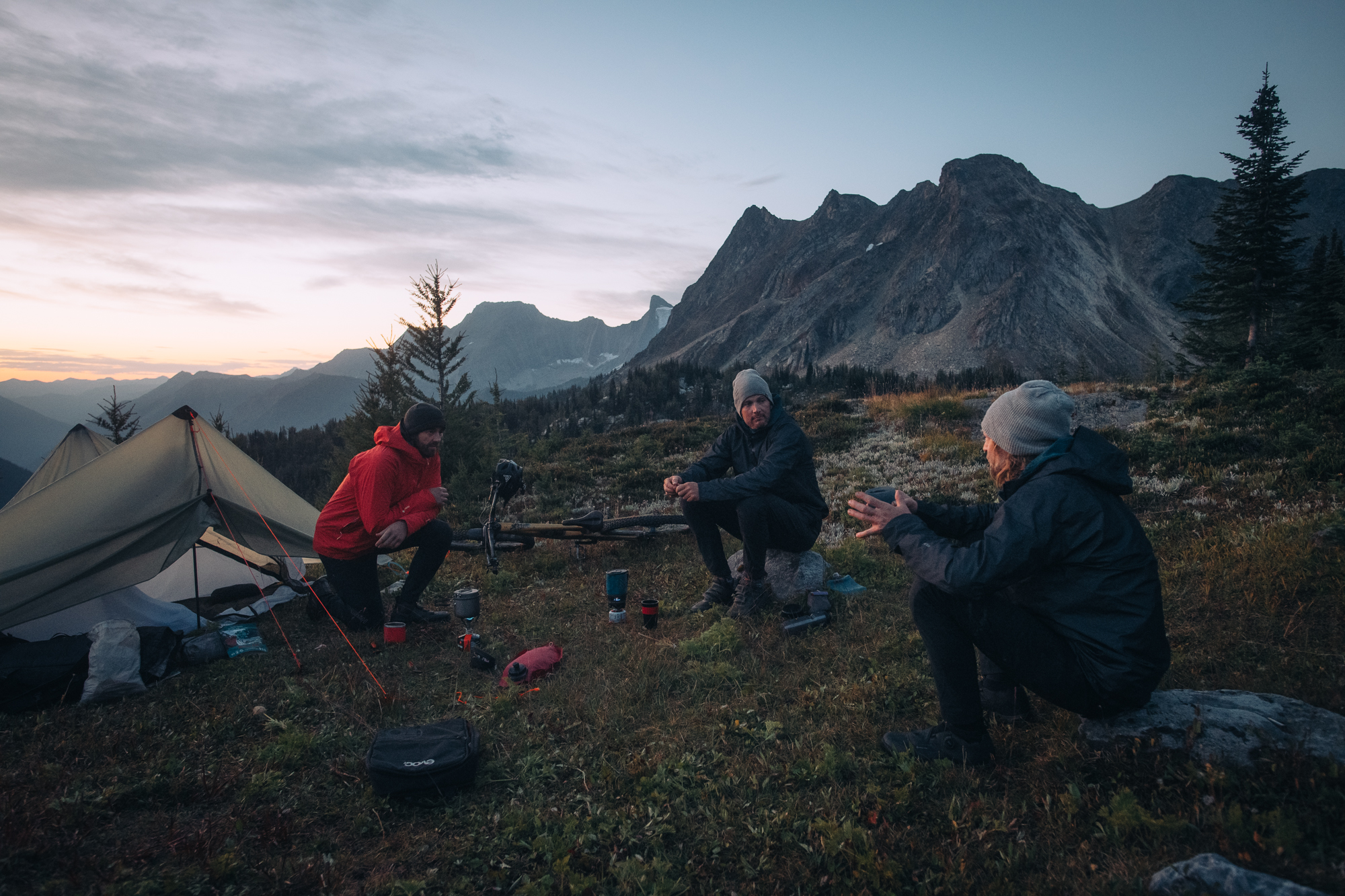 Three riders sitting around stoves at camp below jagged peaks at dusk