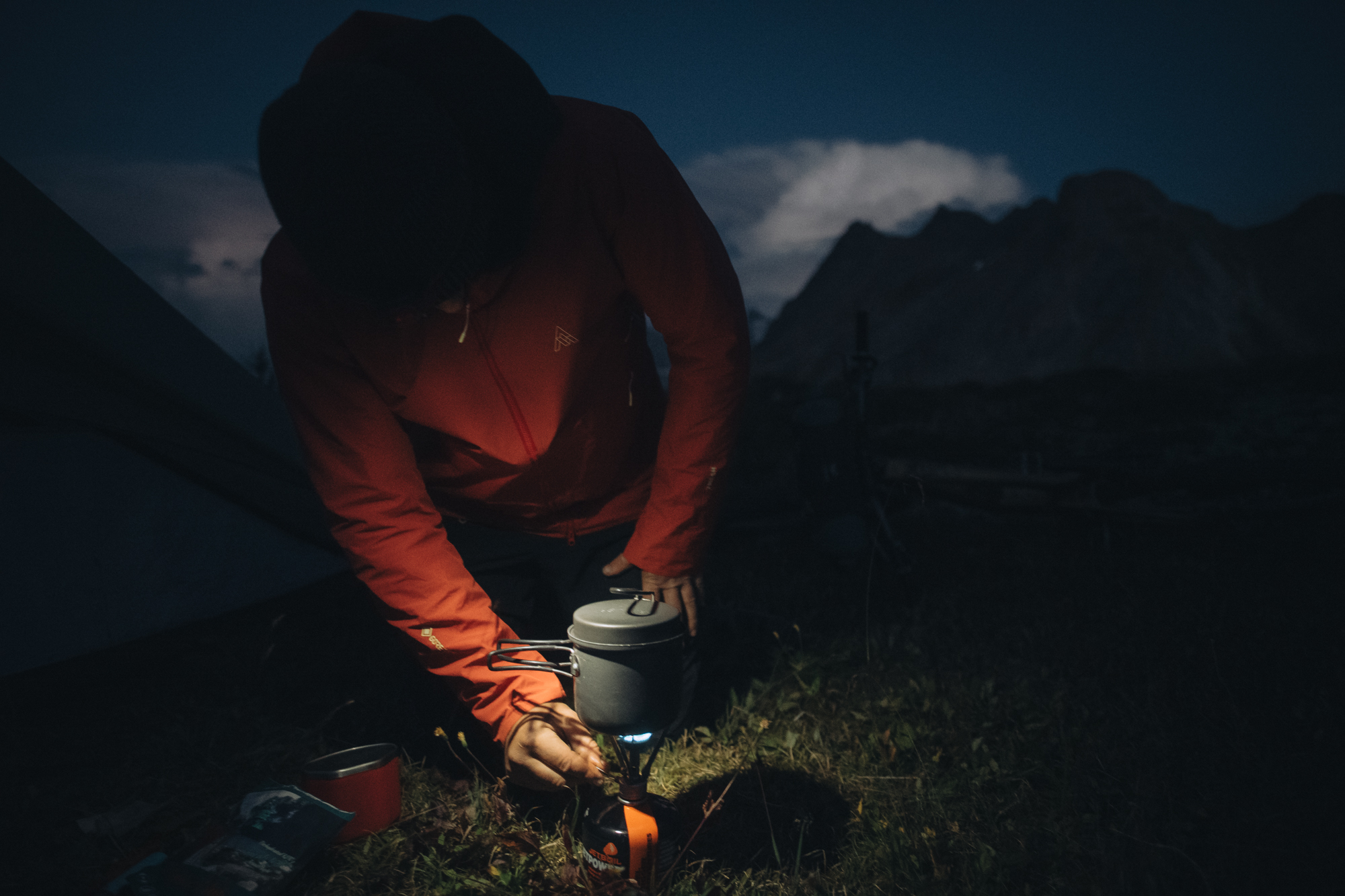 Rider lighting a stove outside a tent under a dark mountain skyline