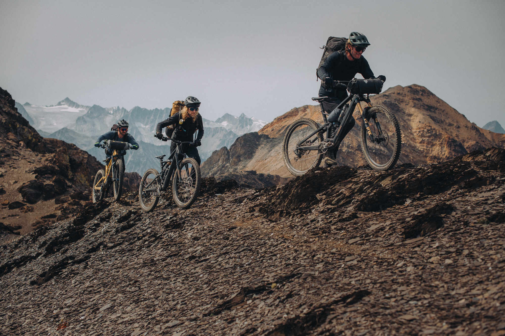 Three riders with loaded bikes on rocky ground below glaciers
