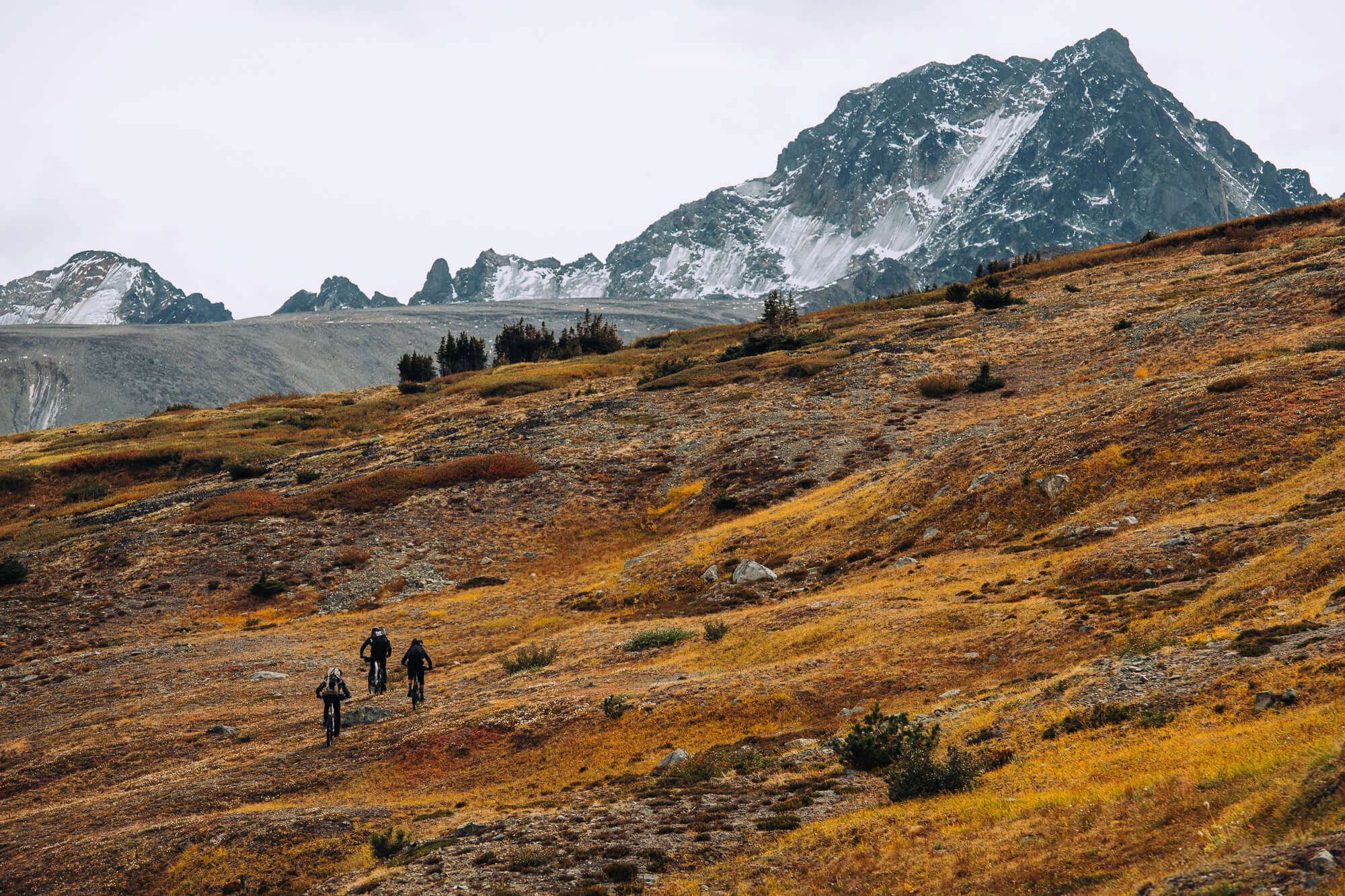 Three riders climbing a golden autumn slope below a snow-streaked peak
