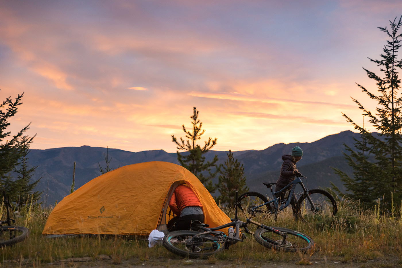 An orange tent pitched at a ridgeline camp under a pink sunset, bikes lying in the grass