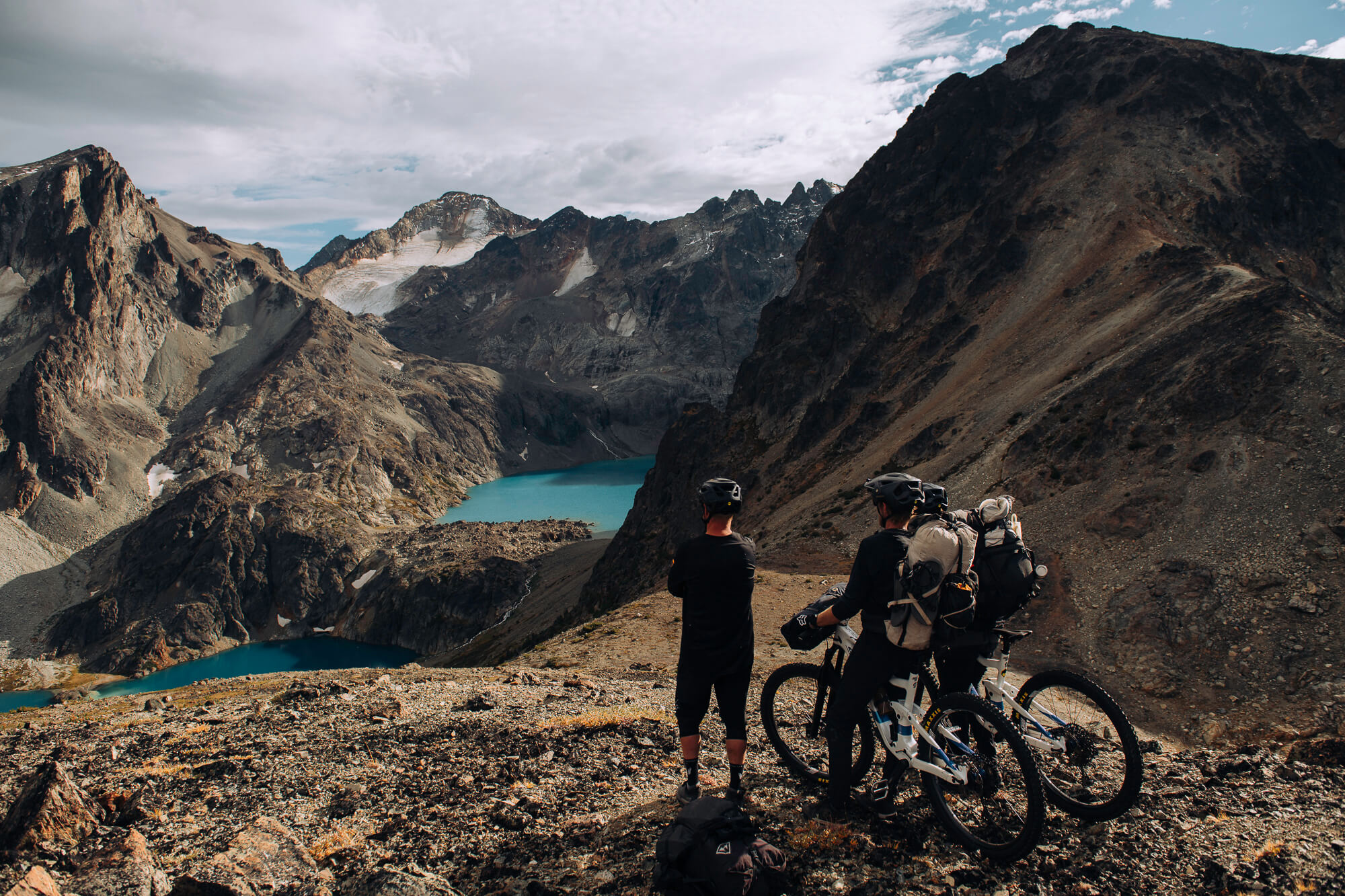 Riders standing with loaded bikes above two turquoise lakes and glaciated peaks