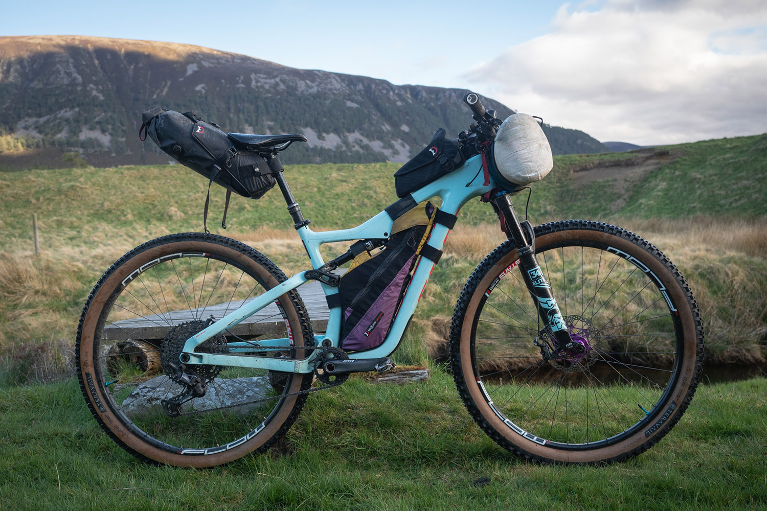 Loaded full-suspension mountain bike with frame bag and seat pack on grass below a hill