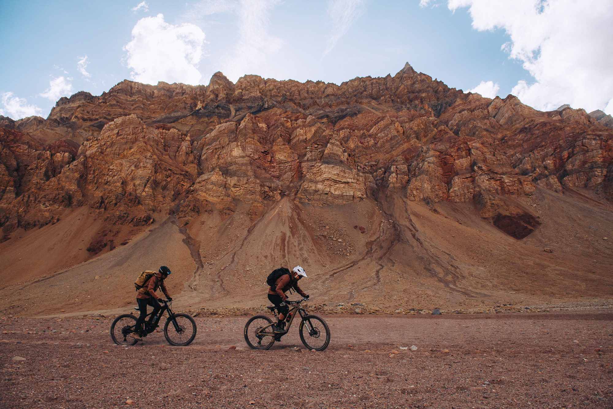 Two riders crossing a wide gravel plain below a red layered rock wall