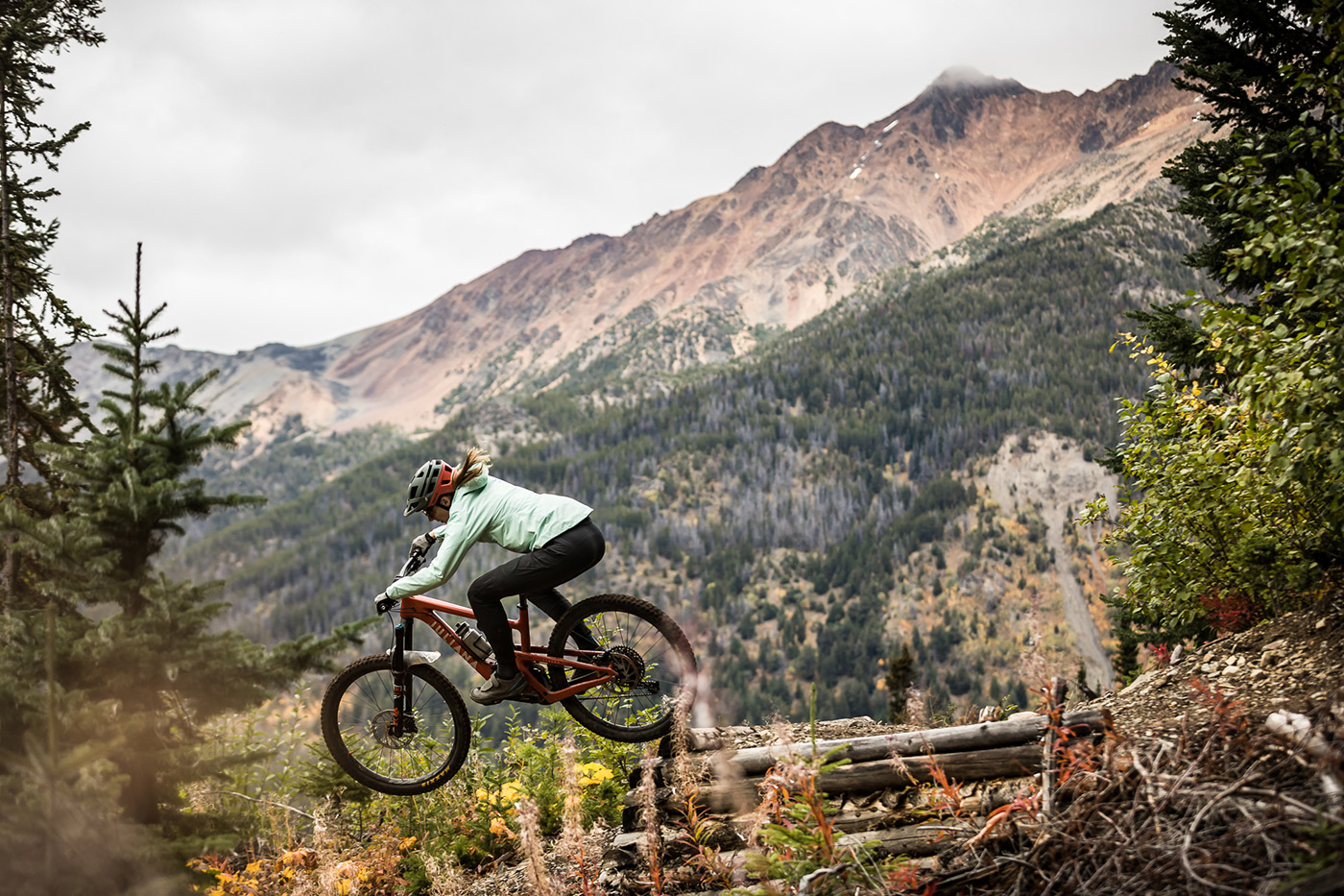 A rider in a mint jacket jumps a trail above a forested valley, red-brown peak behind