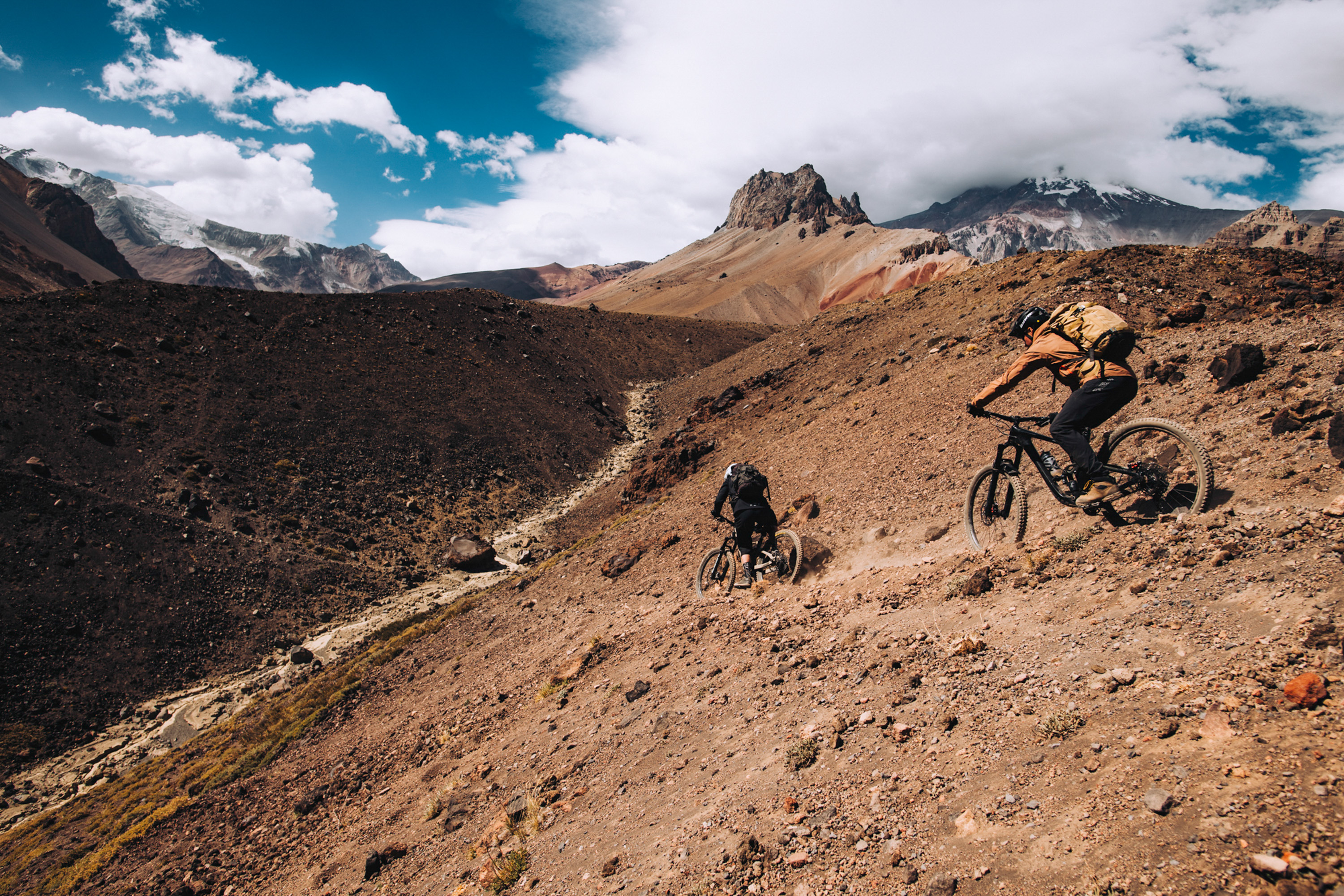 Two riders descending a steep dusty slope below glaciated peaks