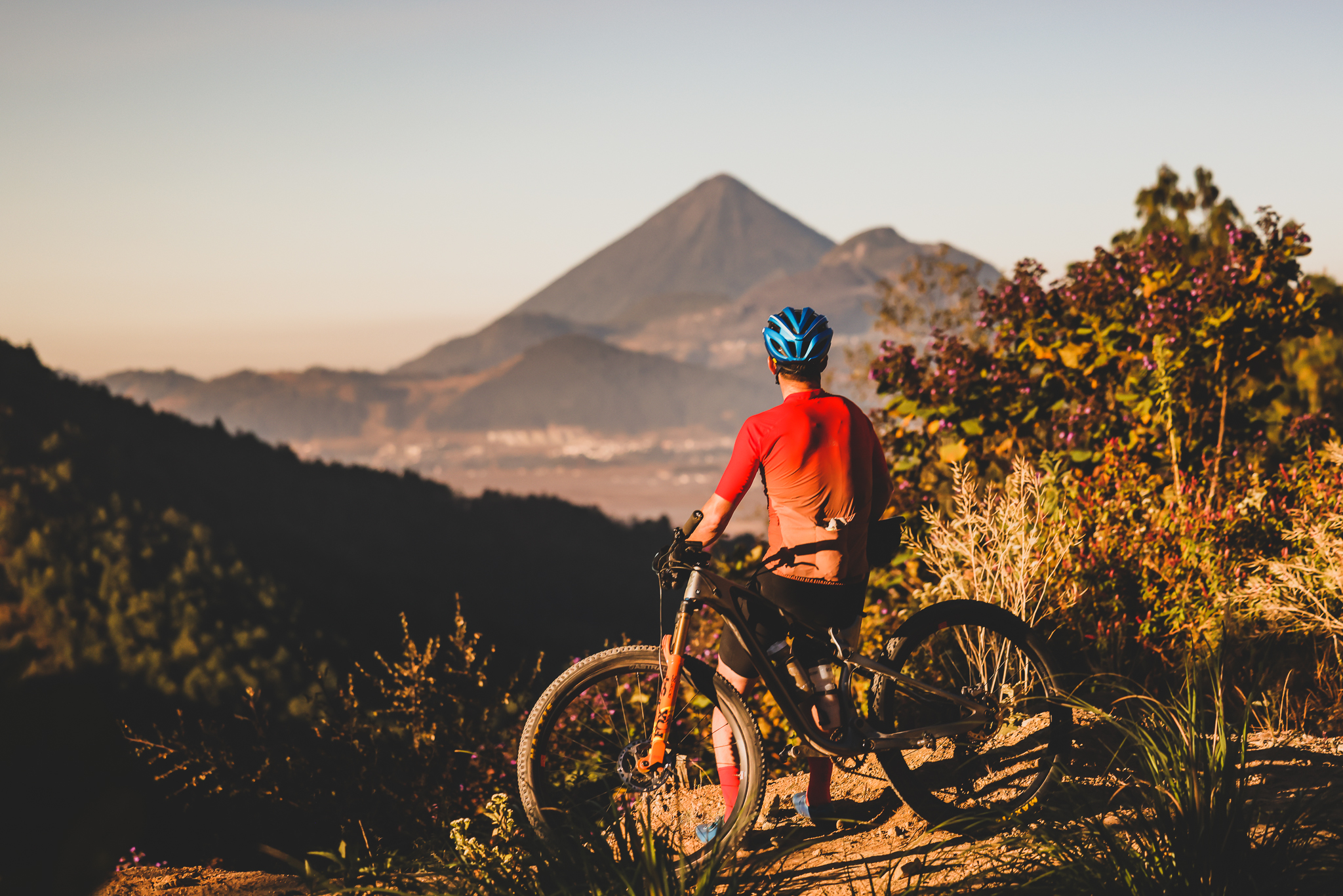 Rider pausing beside a bike on a dirt track facing a distant volcano at sunset