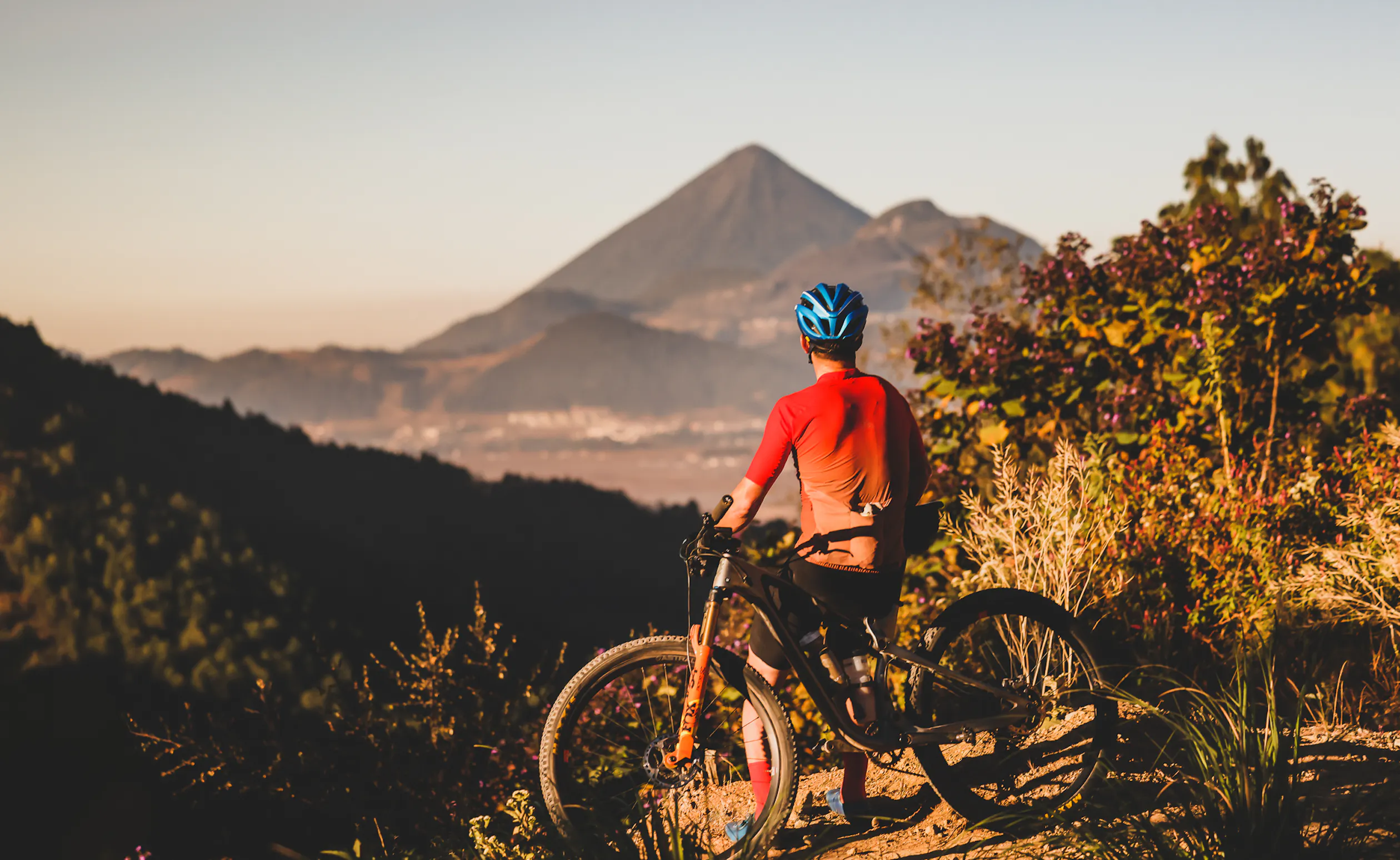 Rider pausing beside a bike on a dirt track facing a distant volcano at sunset