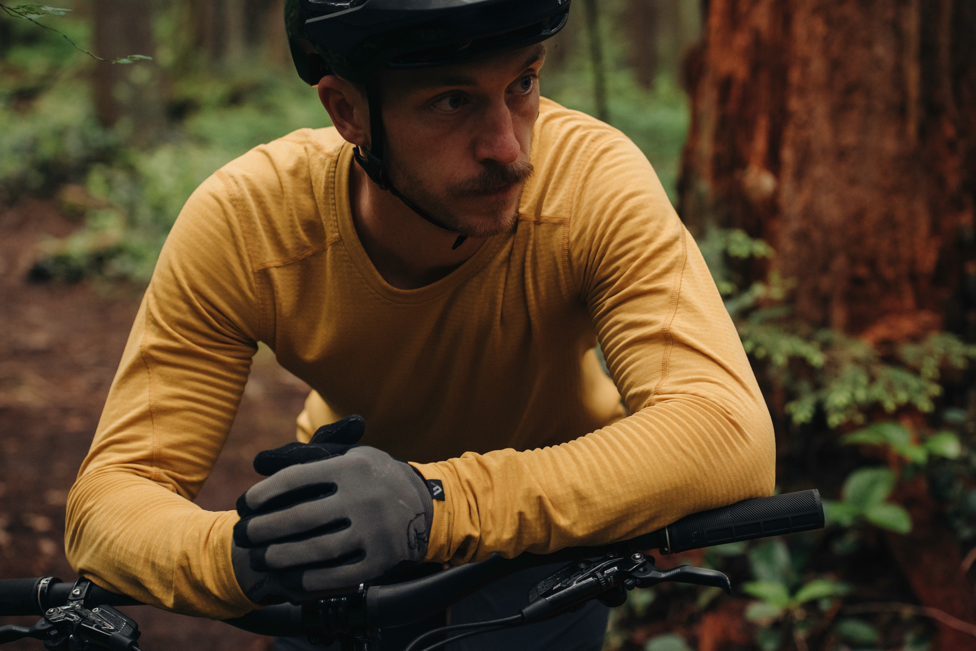 Rider in a mustard merino base layer leaning on mountain bike bars in the forest