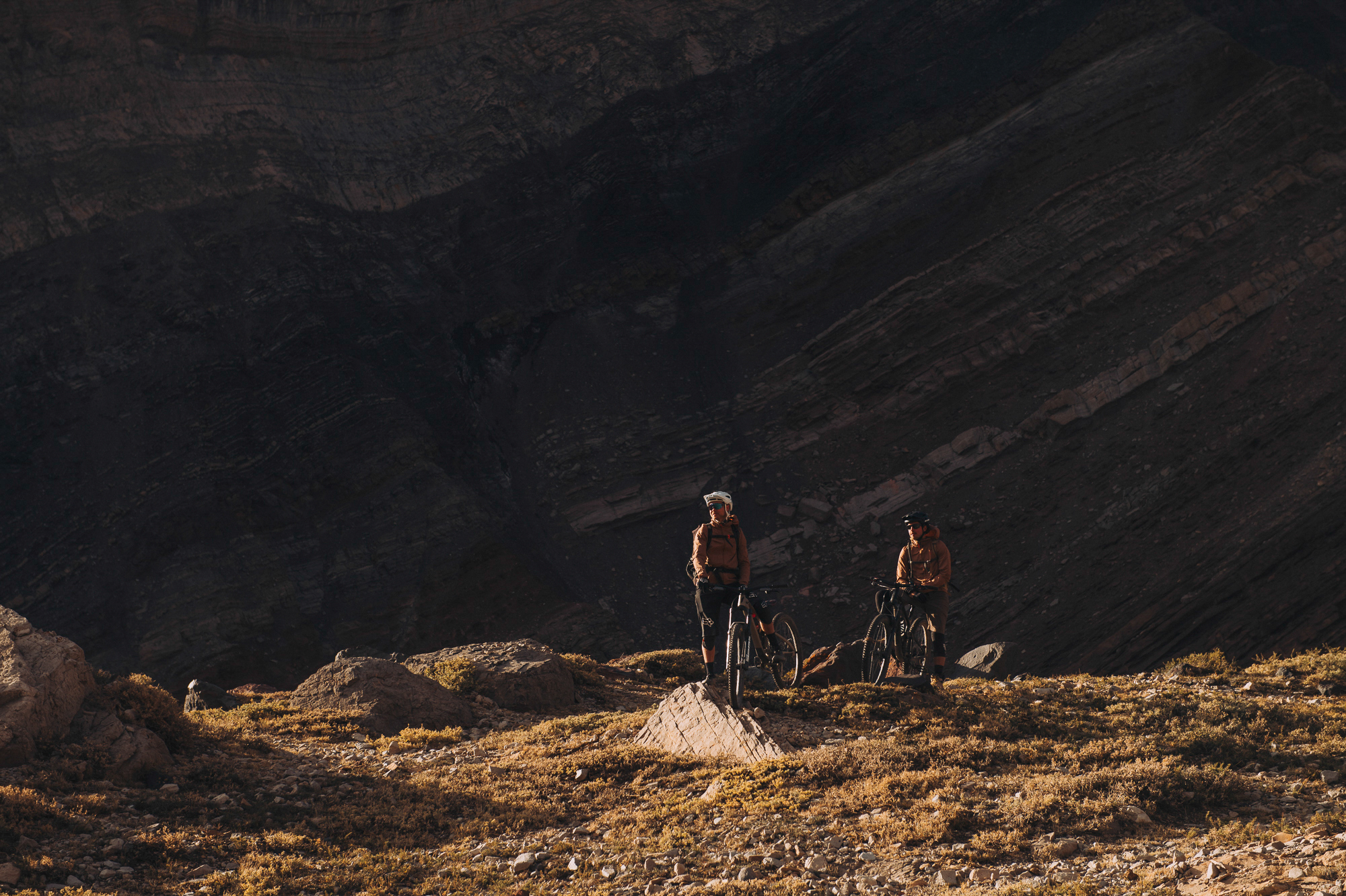 Two riders standing with bikes on a grassy bench under a vast dark wall