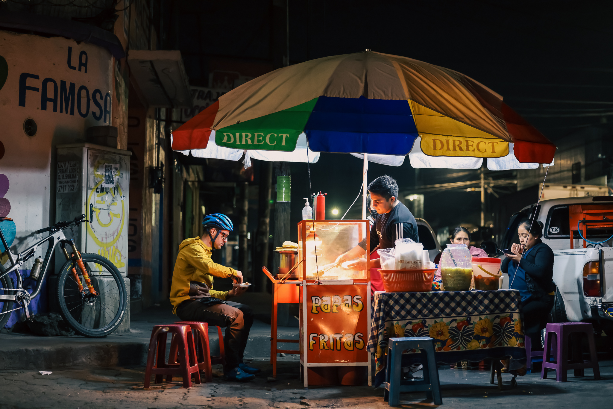 Rider eating at a lit street food stall under a striped umbrella at night