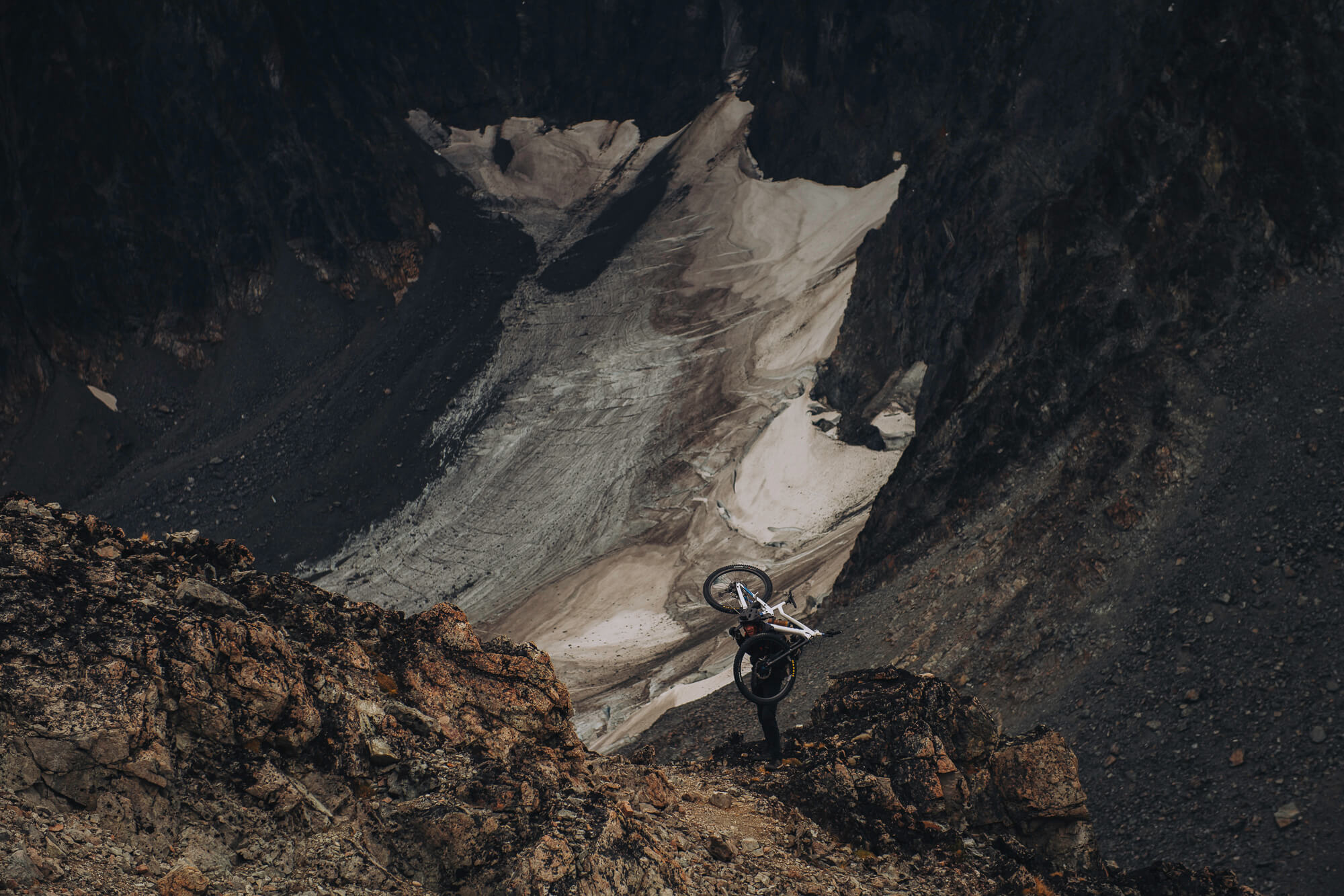 Rider carrying a bike up a rocky slope above a dirt-streaked glacier
