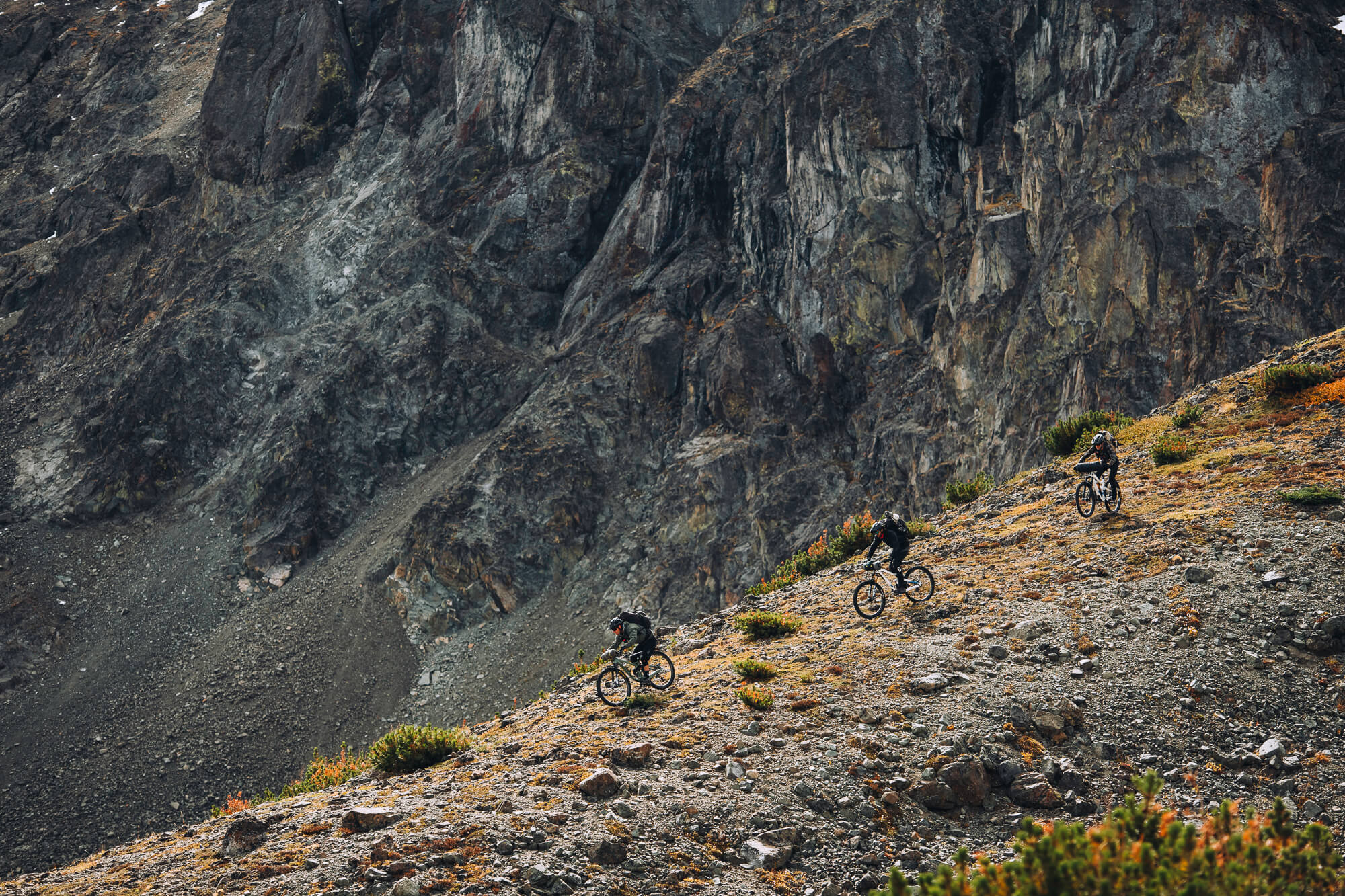 Three riders descending a loose slope below a large rock face