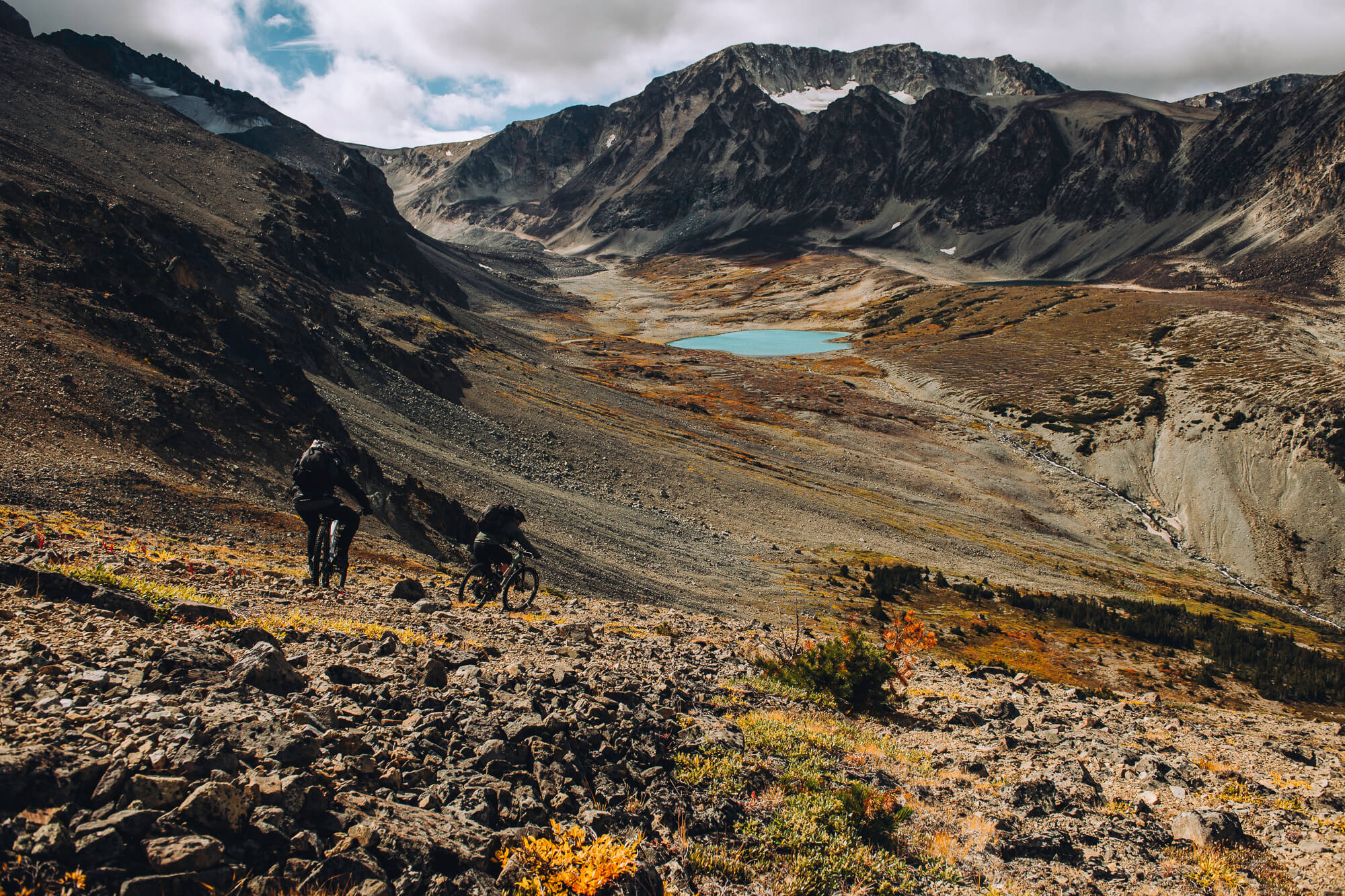 Two riders descending a scree slope toward a turquoise alpine lake