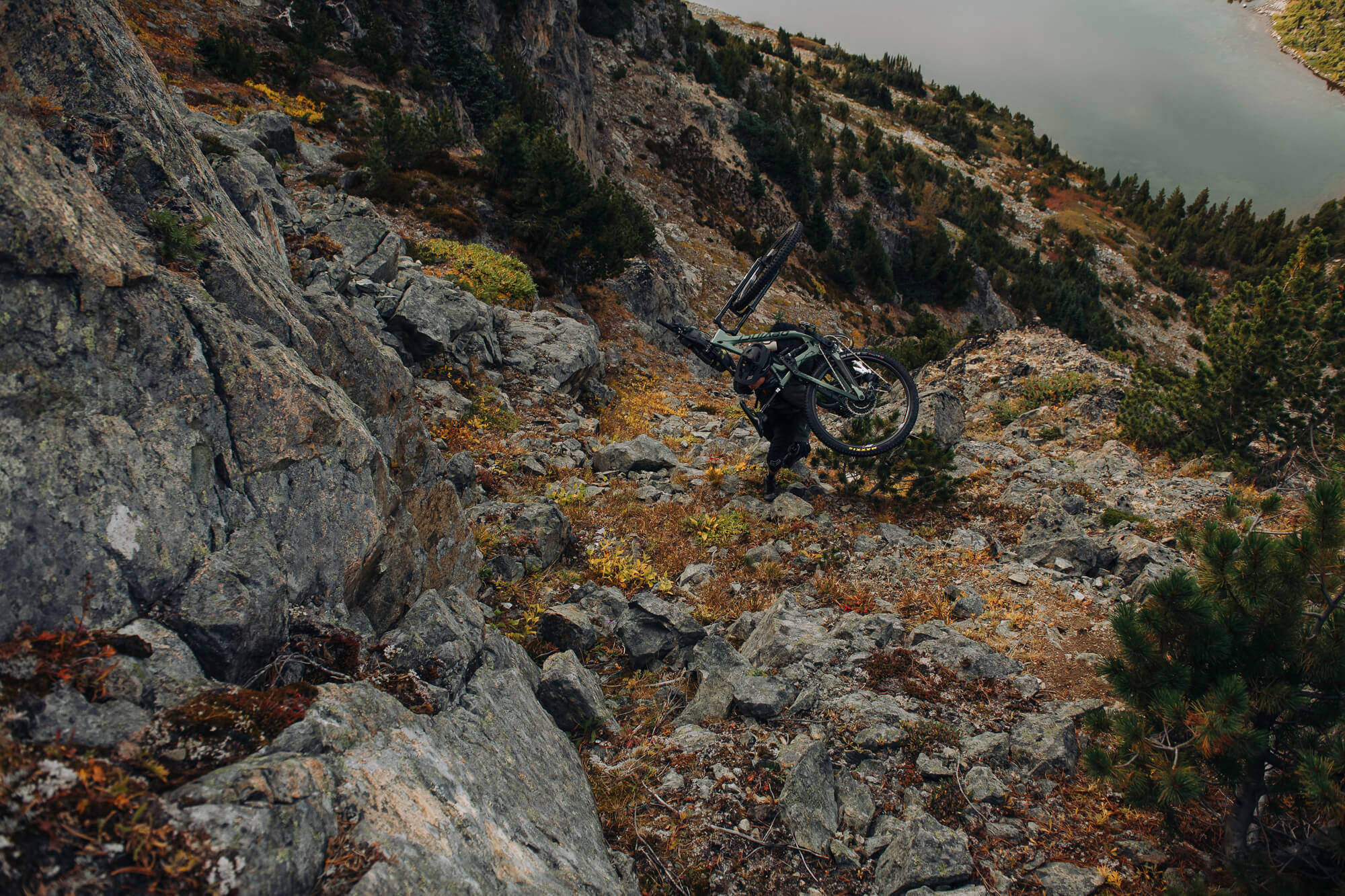 Rider carrying a mountain bike on one shoulder down a steep boulder slope above a lake