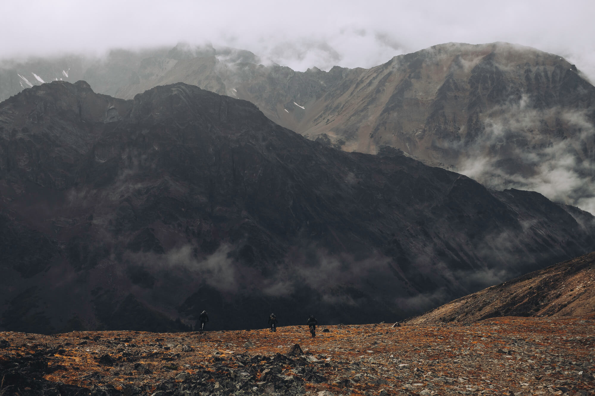 Three riders crossing orange alpine tundra below cloud-covered mountains